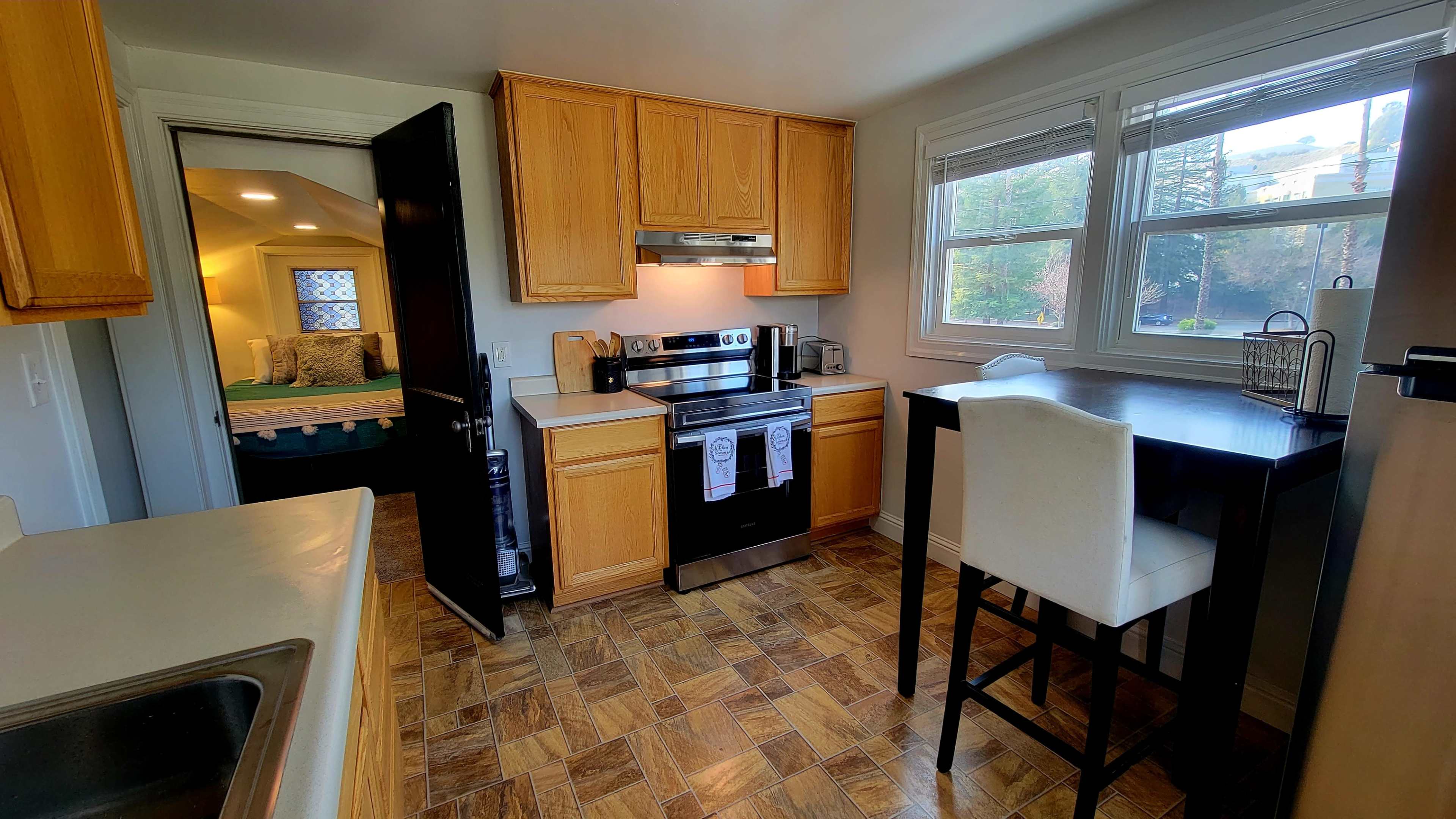 The image shows a modern kitchen with wooden cabinets, an oven, and a small dining table near the windows.