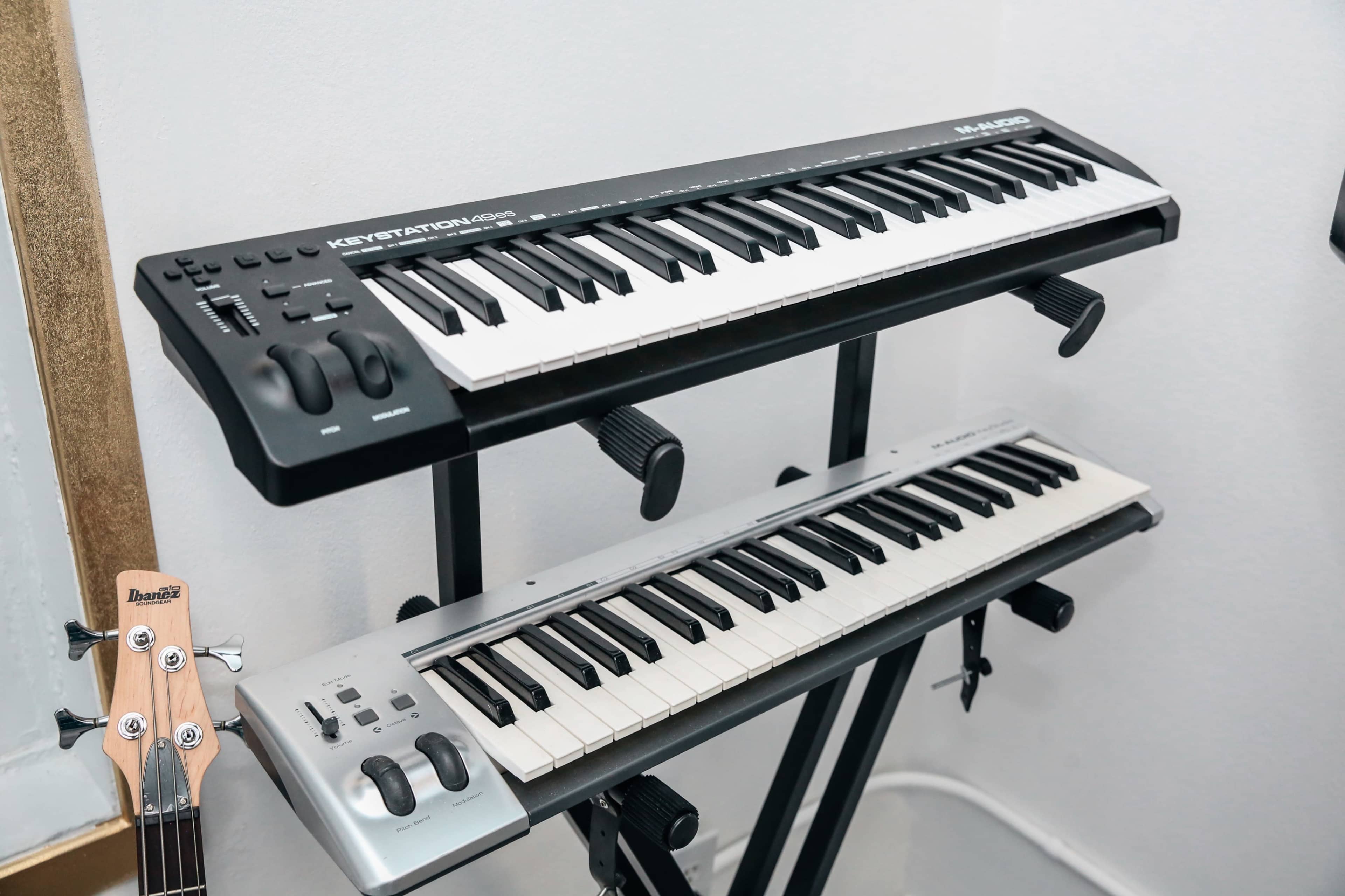 The image shows two keyboard instruments arranged on a music stand, with a bass guitar partially visible to the left.