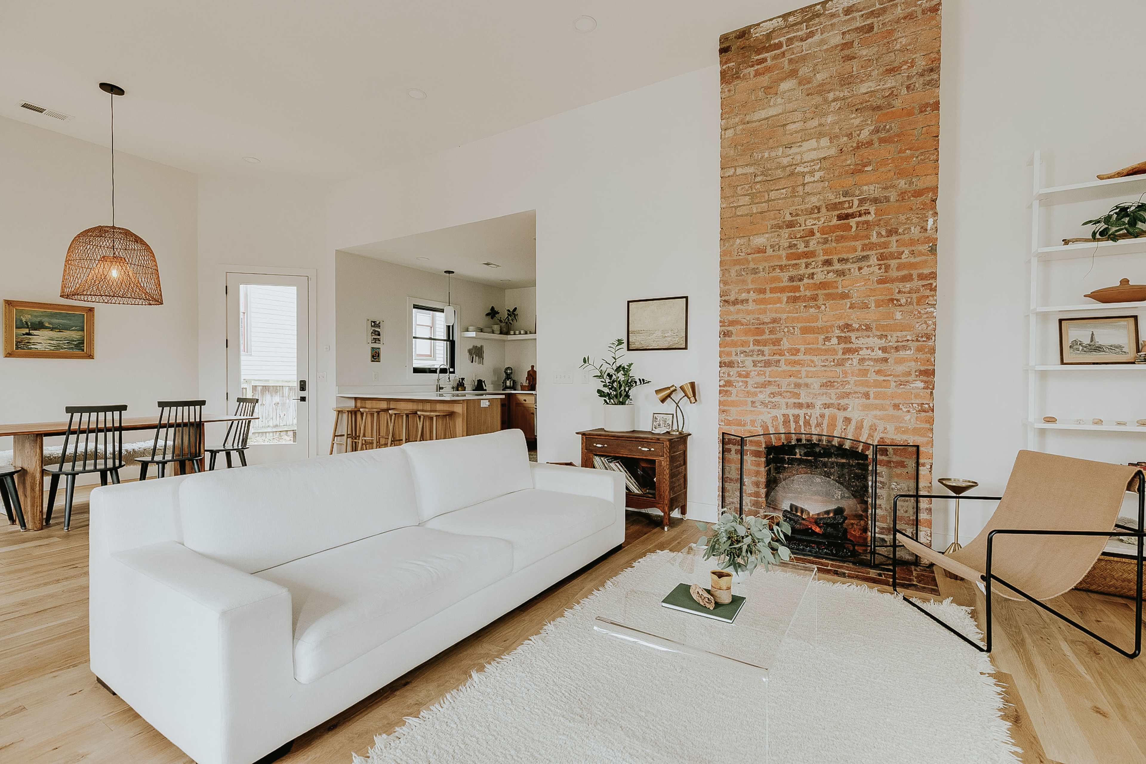 A modern living room features a white sofa, a brick accent wall with a fireplace, and a decorative rug, alongside a dining area visible through an open space.
