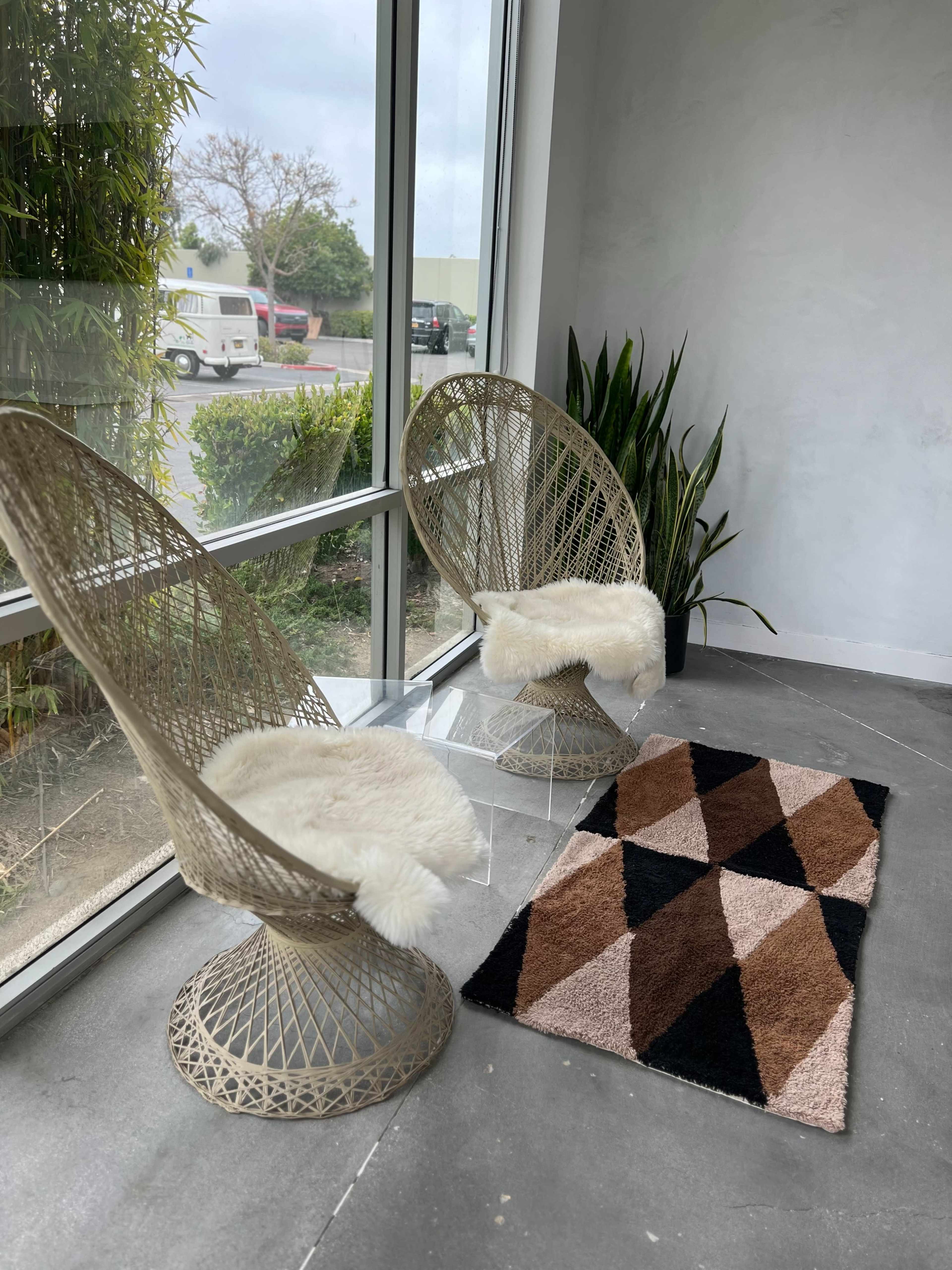 Two woven chairs with fur throws are positioned next to a geometric patterned rug on a concrete floor beside large windows.