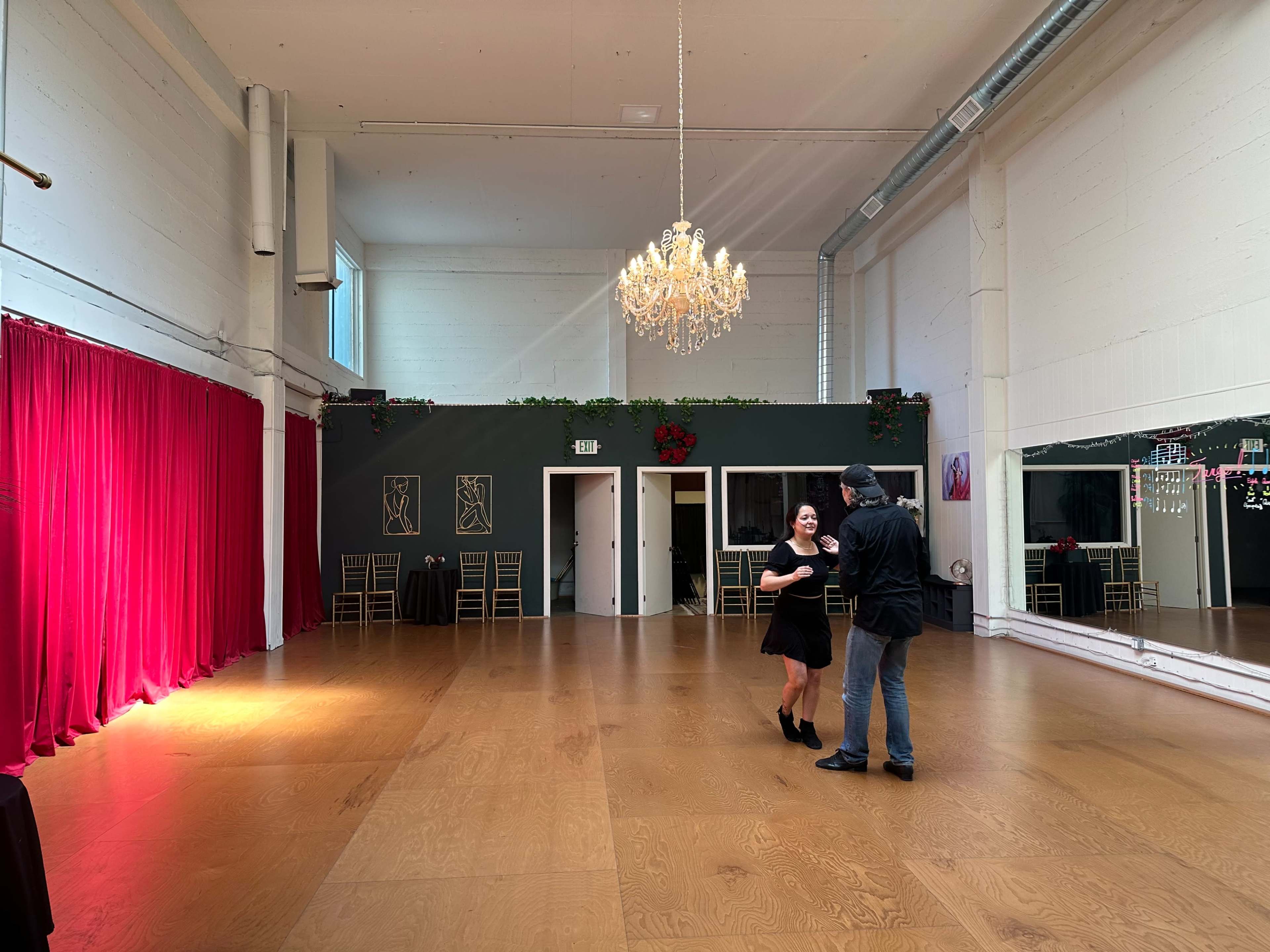 A couple dances in a spacious, well-lit room with a chandelier and red curtains.