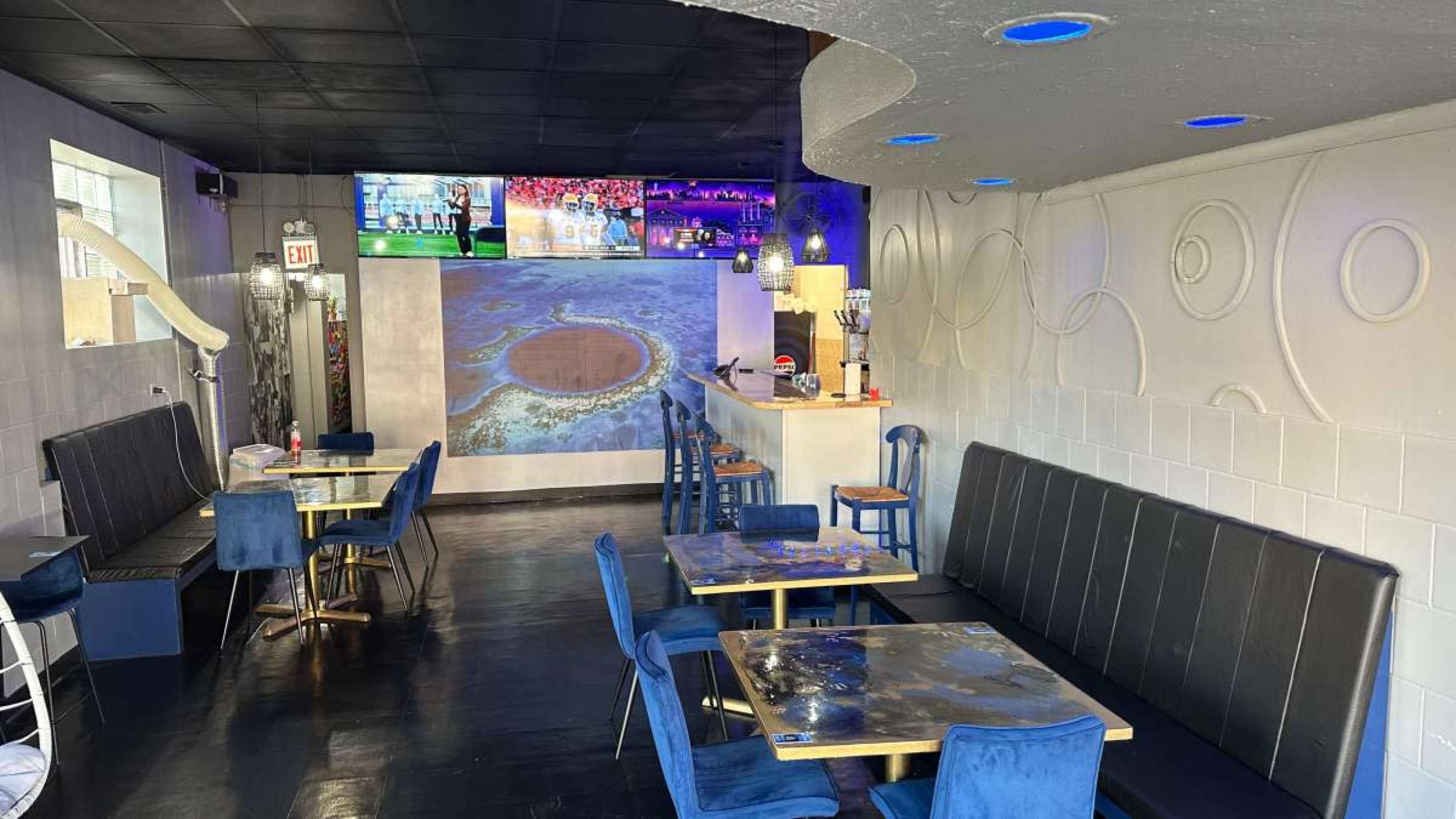 The image shows a modern restaurant interior with blue velvet chairs, tables featuring reflective surfaces, and a large screen displaying a visual on the wall.