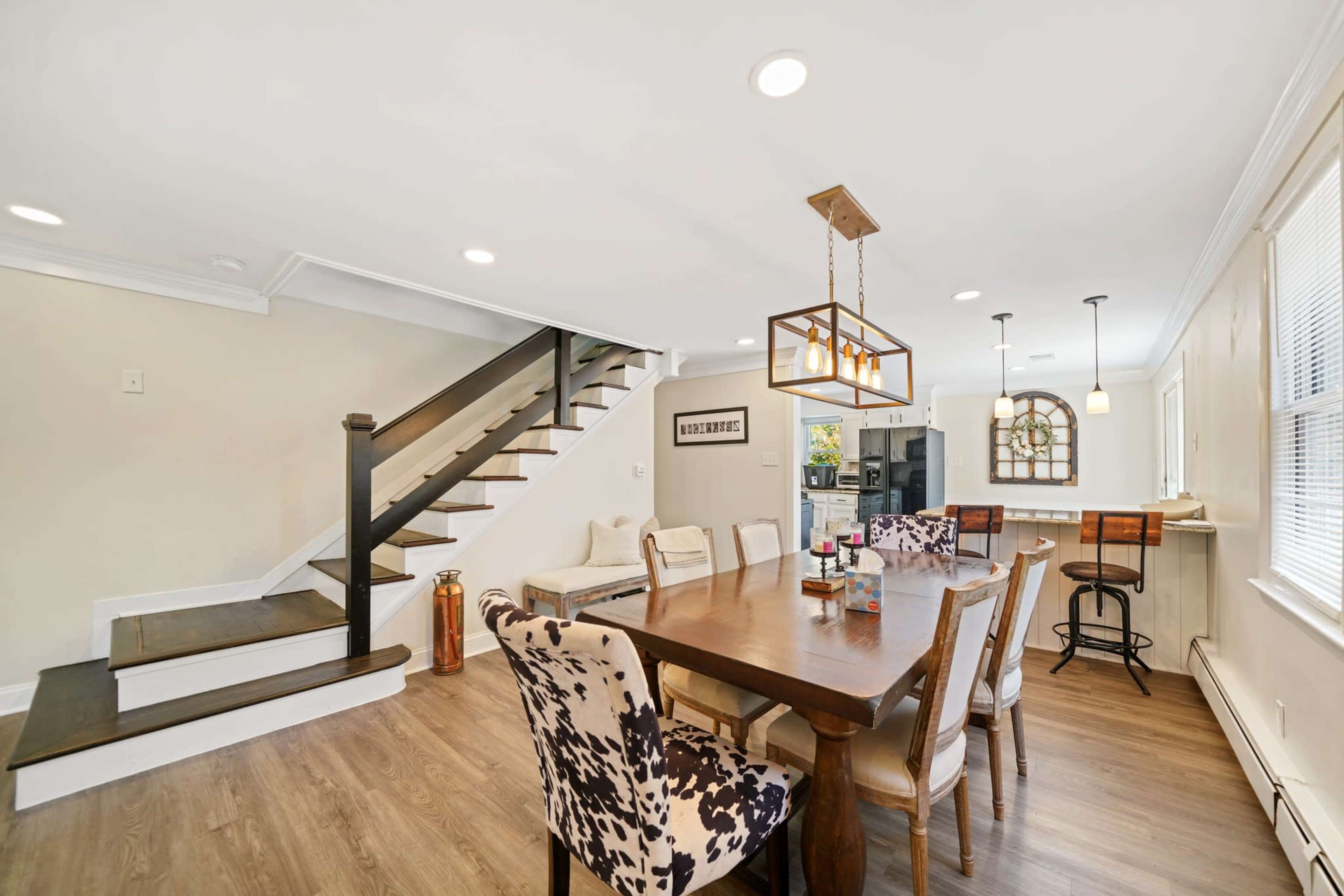 A dining area with a wooden table surrounded by patterned chairs, adjacent to a staircase and a kitchen area with bar seating.