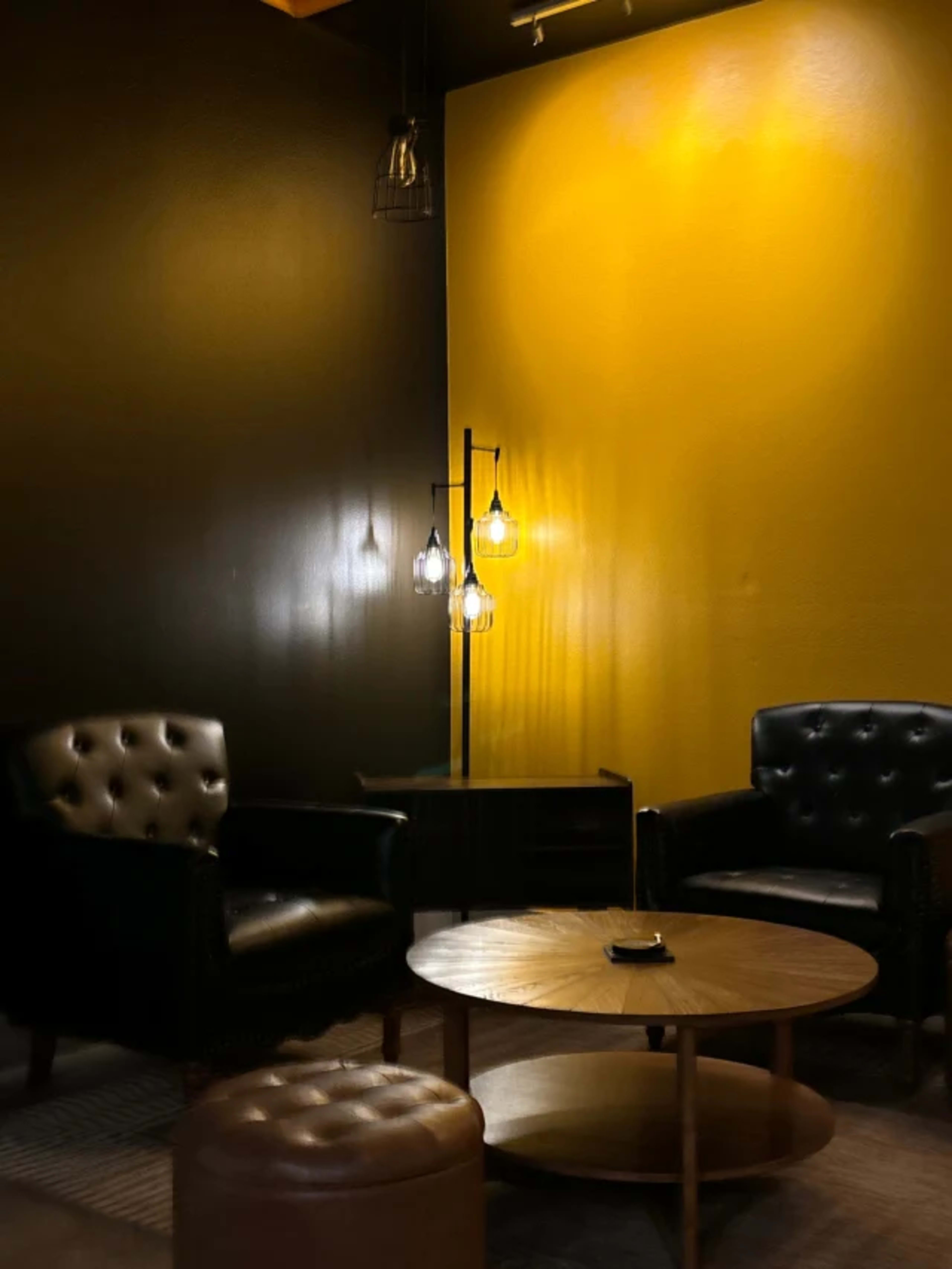 The image depicts a cozy seating area with two black leather chairs facing a wooden coffee table, illuminated by pendant lights on a yellow and black wall.