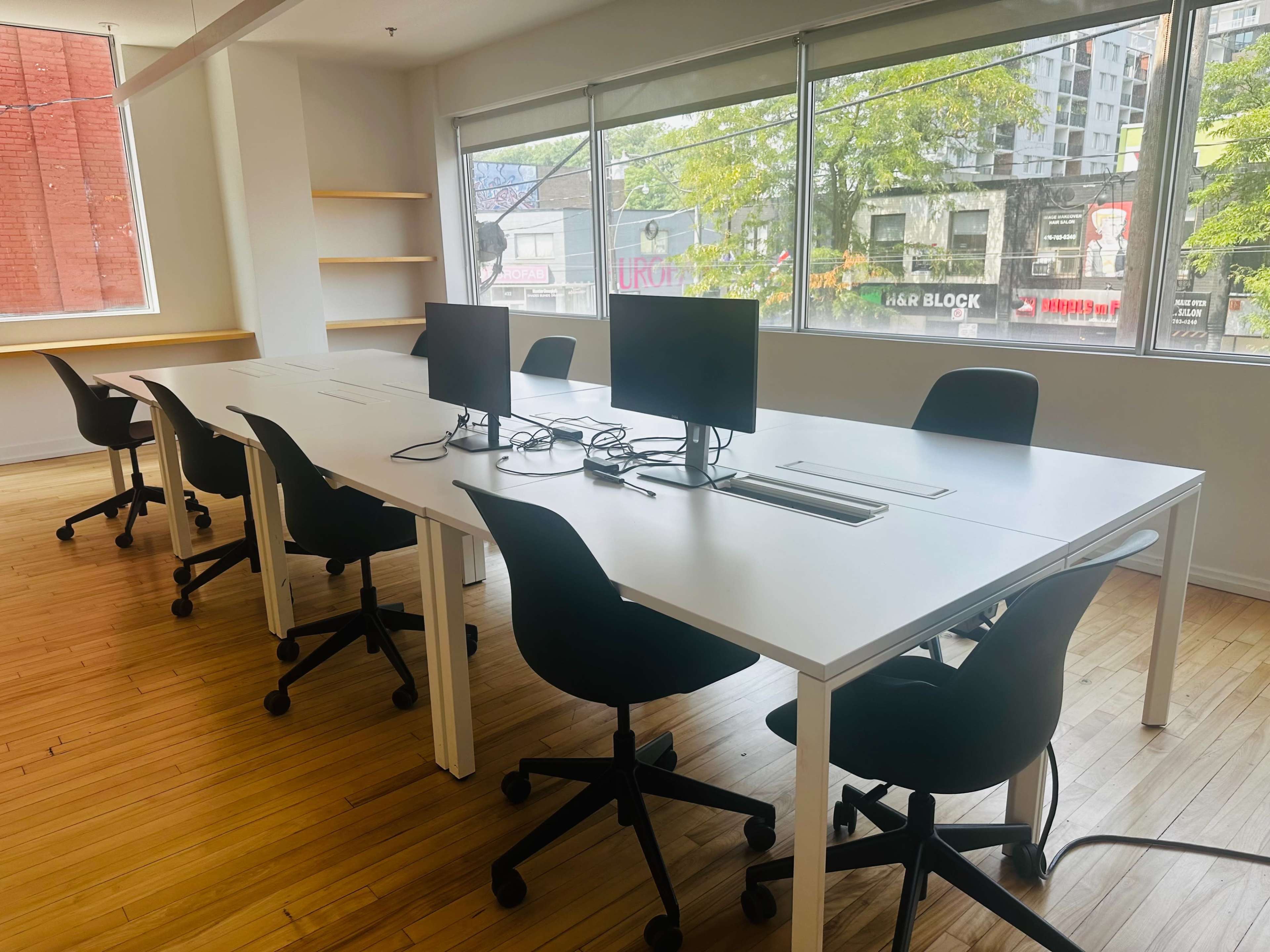 A modern office space features a long white table with multiple computer monitors and black swivel chairs, illuminated by large windows.