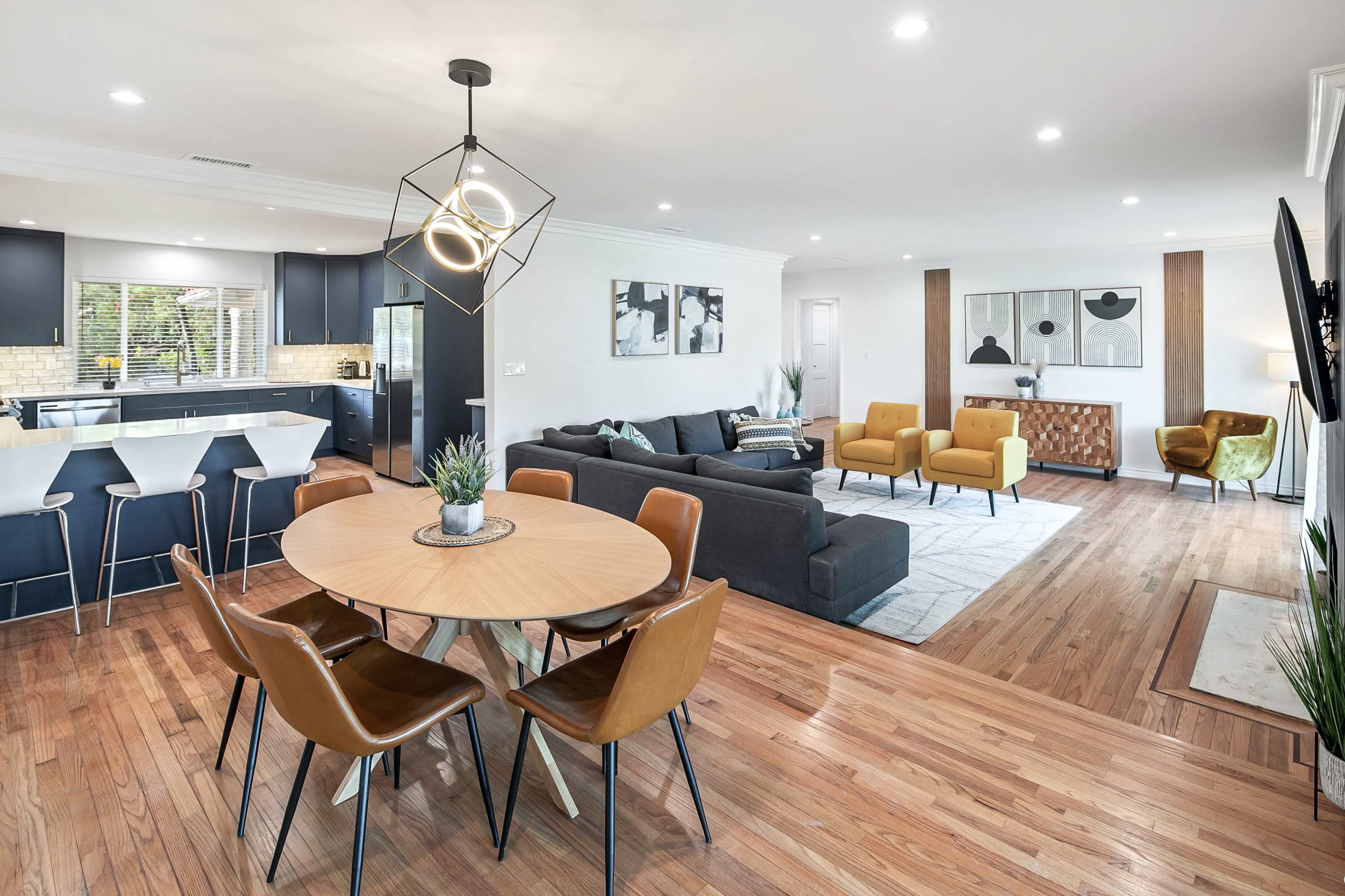 The image shows an open-concept living and dining area featuring a round dining table with chairs, a modern kitchen with dark cabinetry, and a cozy seating arrangement with a sofa and armchairs.