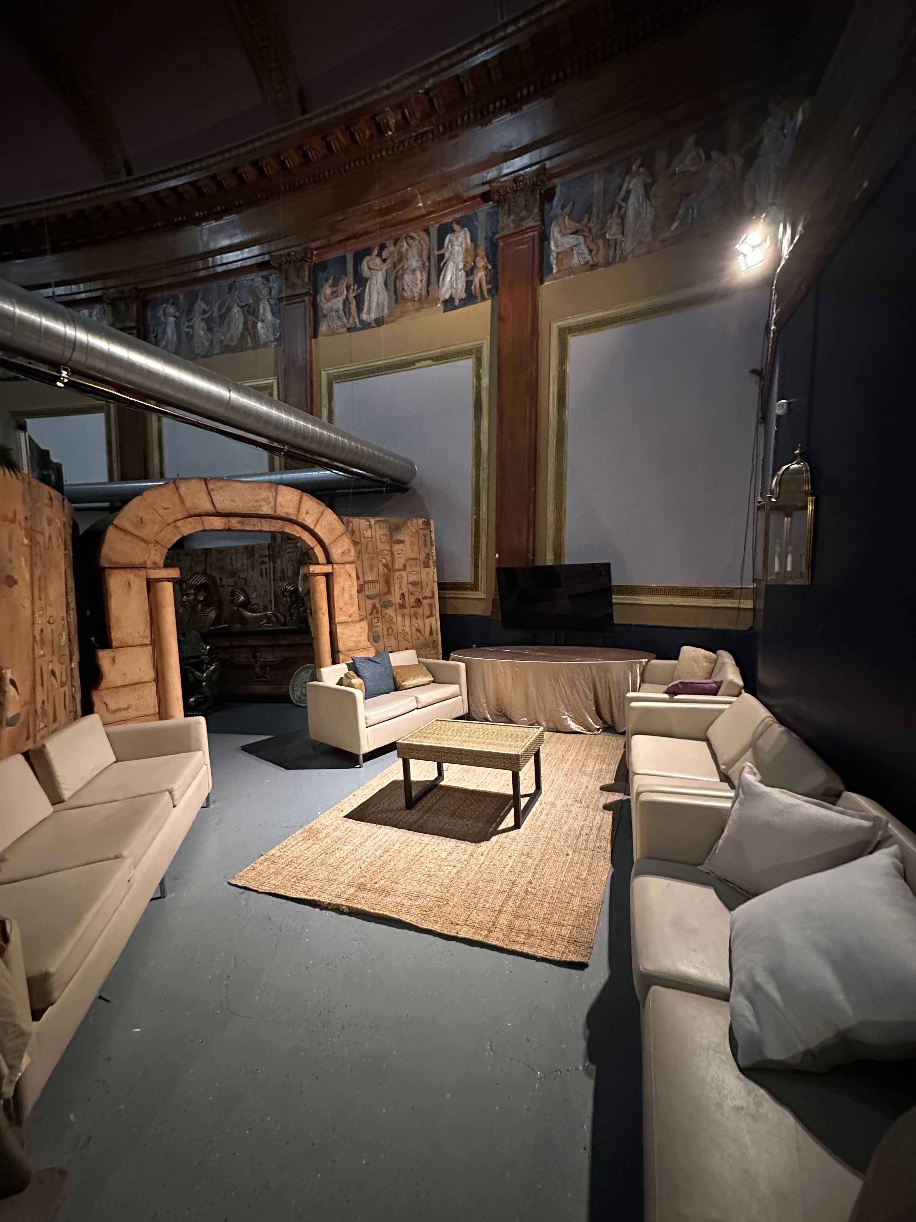 The image shows a spacious room with a circular seating arrangement, featuring modern sofas, a central coffee table, and decorative wall murals.