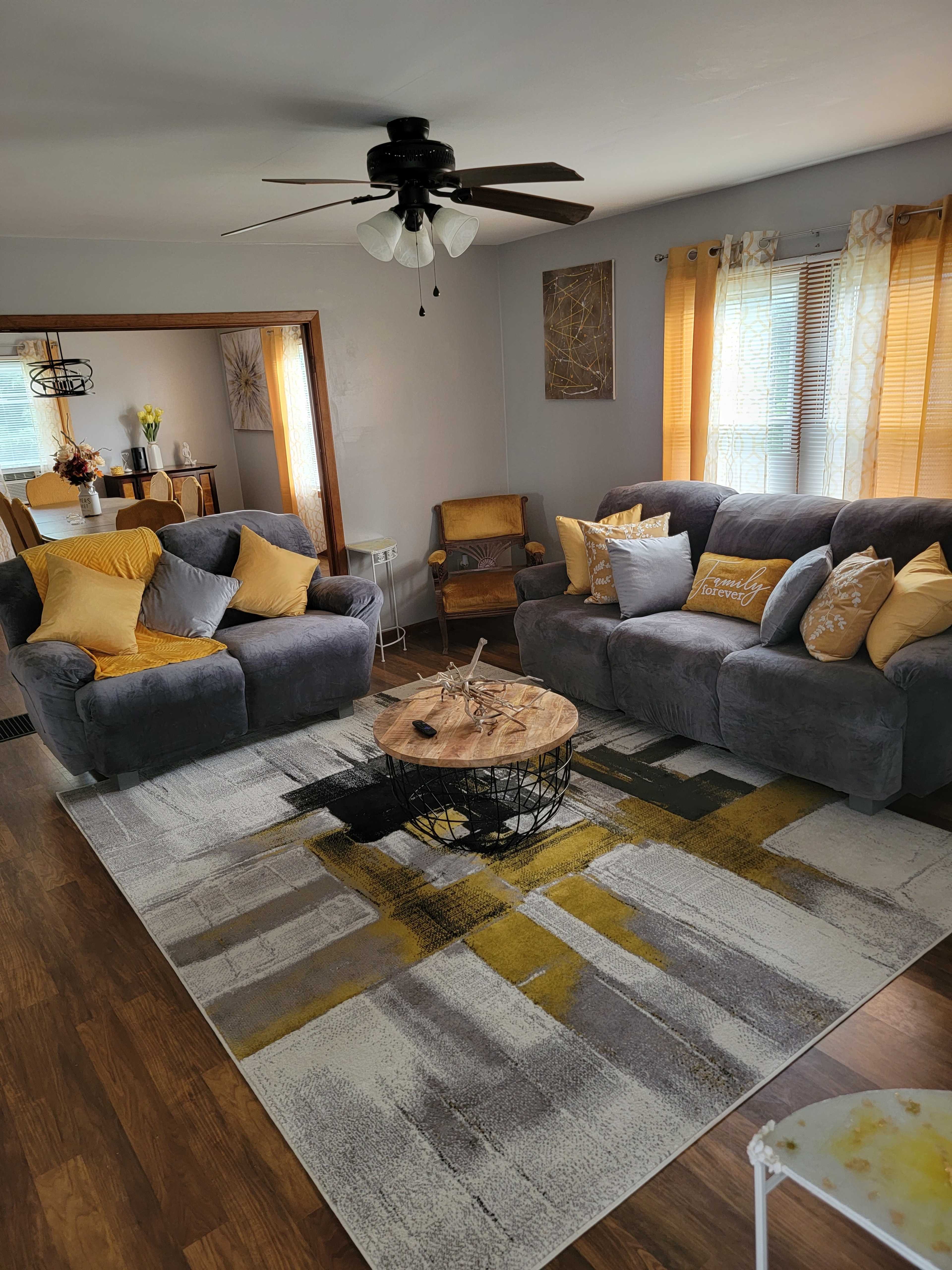 The living room features two gray couches, a circular wooden coffee table, and a patterned rug, with natural light streaming through the window shades.