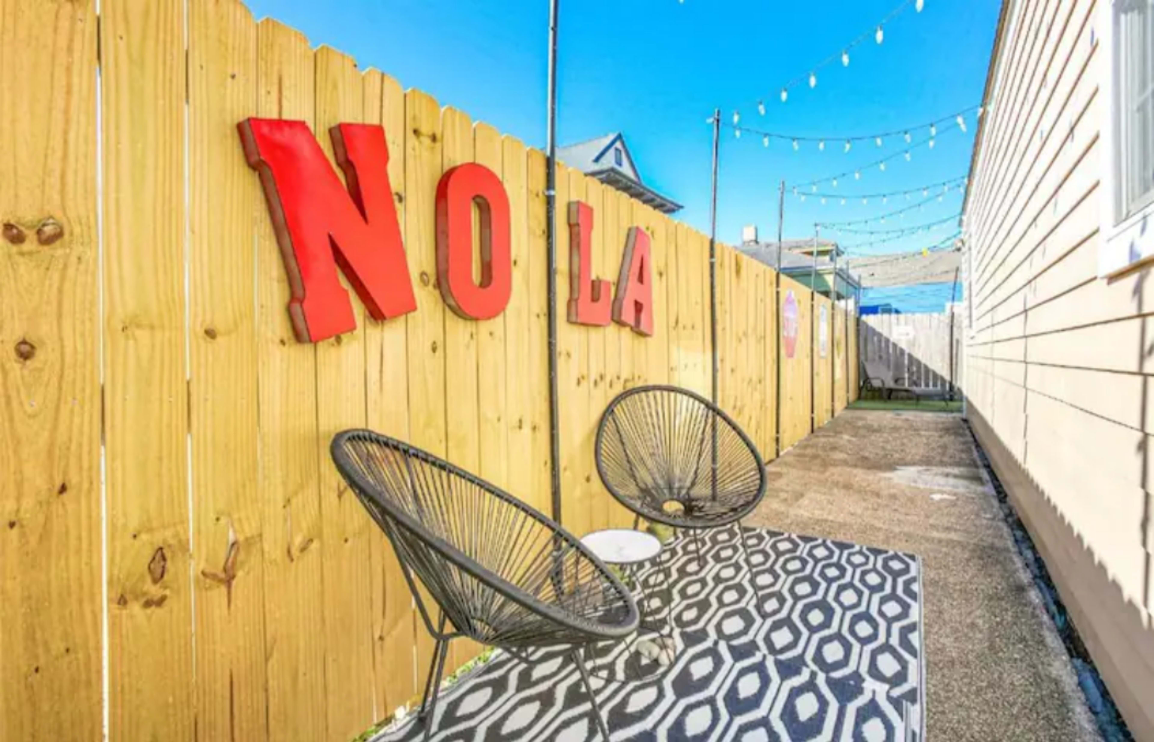 A narrow outdoor area with a wooden fence displaying the letters "NOLA," two black wire chairs, and a patterned rug under a clear blue sky.