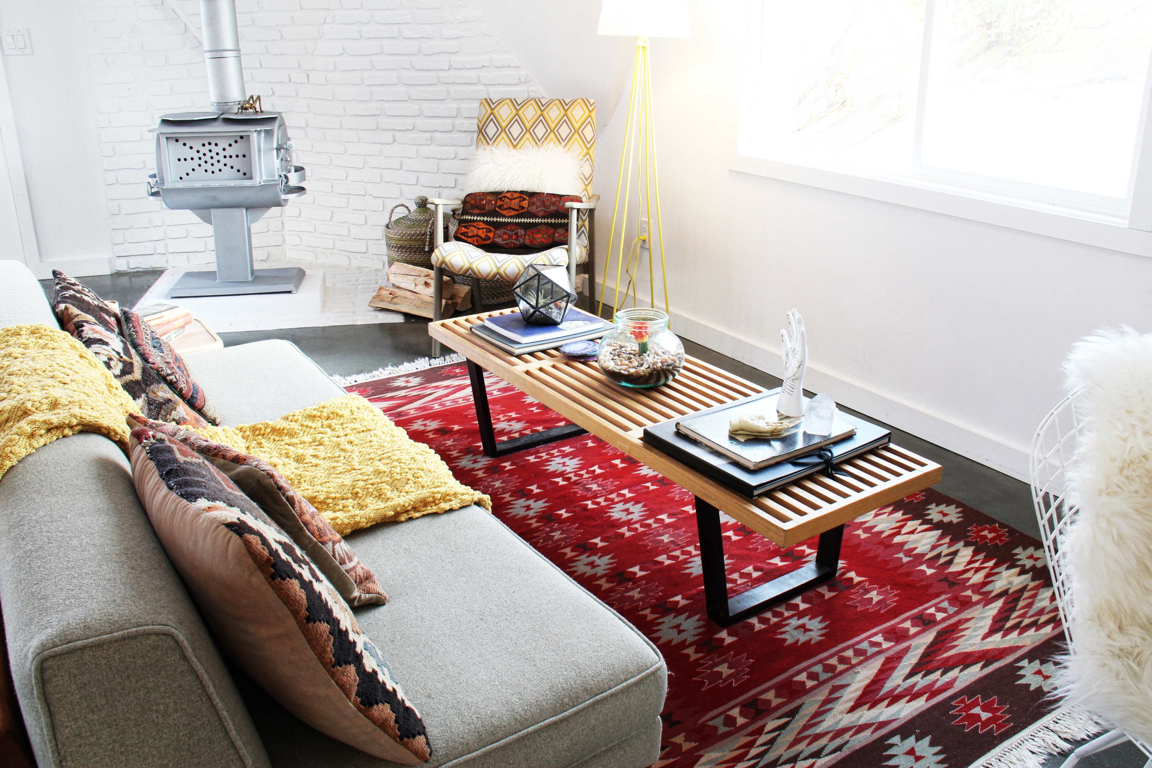 The image shows a modern living room with a gray sofa, a wooden coffee table with slatted design, and a patterned red rug.