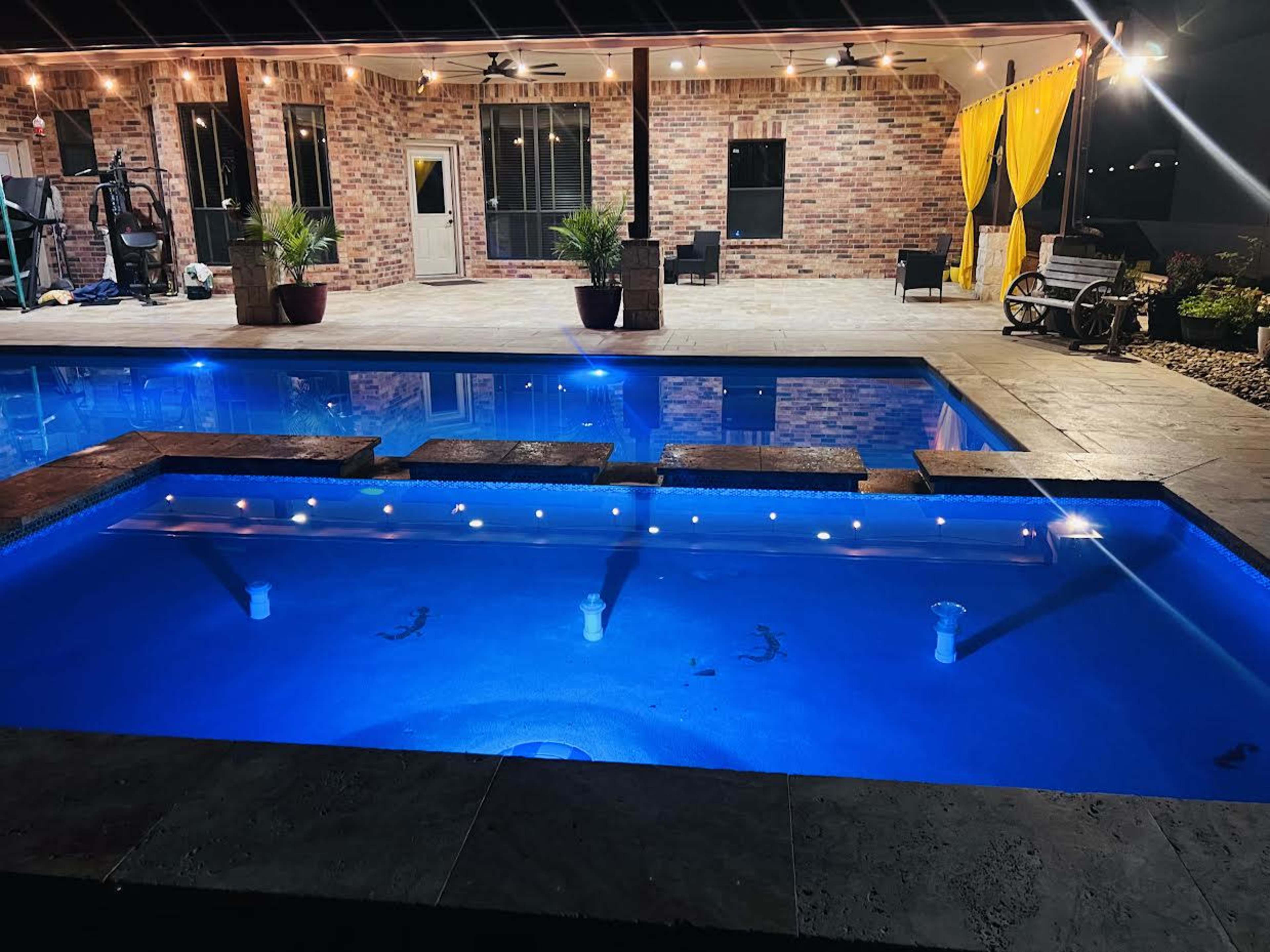 The image shows a well-lit outdoor pool area with a series of steps leading into the water and a brick house in the background.