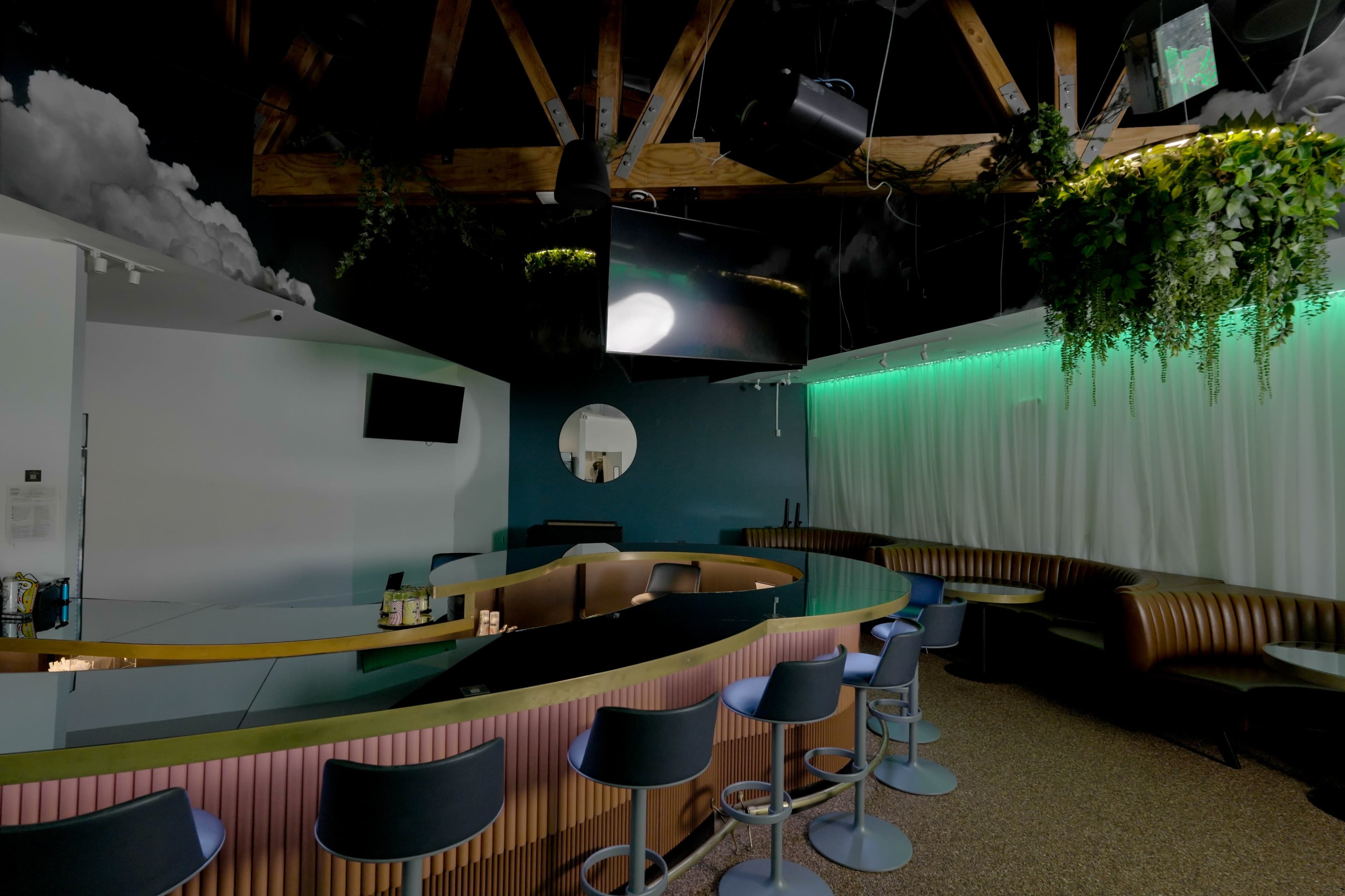 The image shows a modern bar interior featuring a curved counter, high stools, and a backdrop with green ambient lighting and lush greenery.