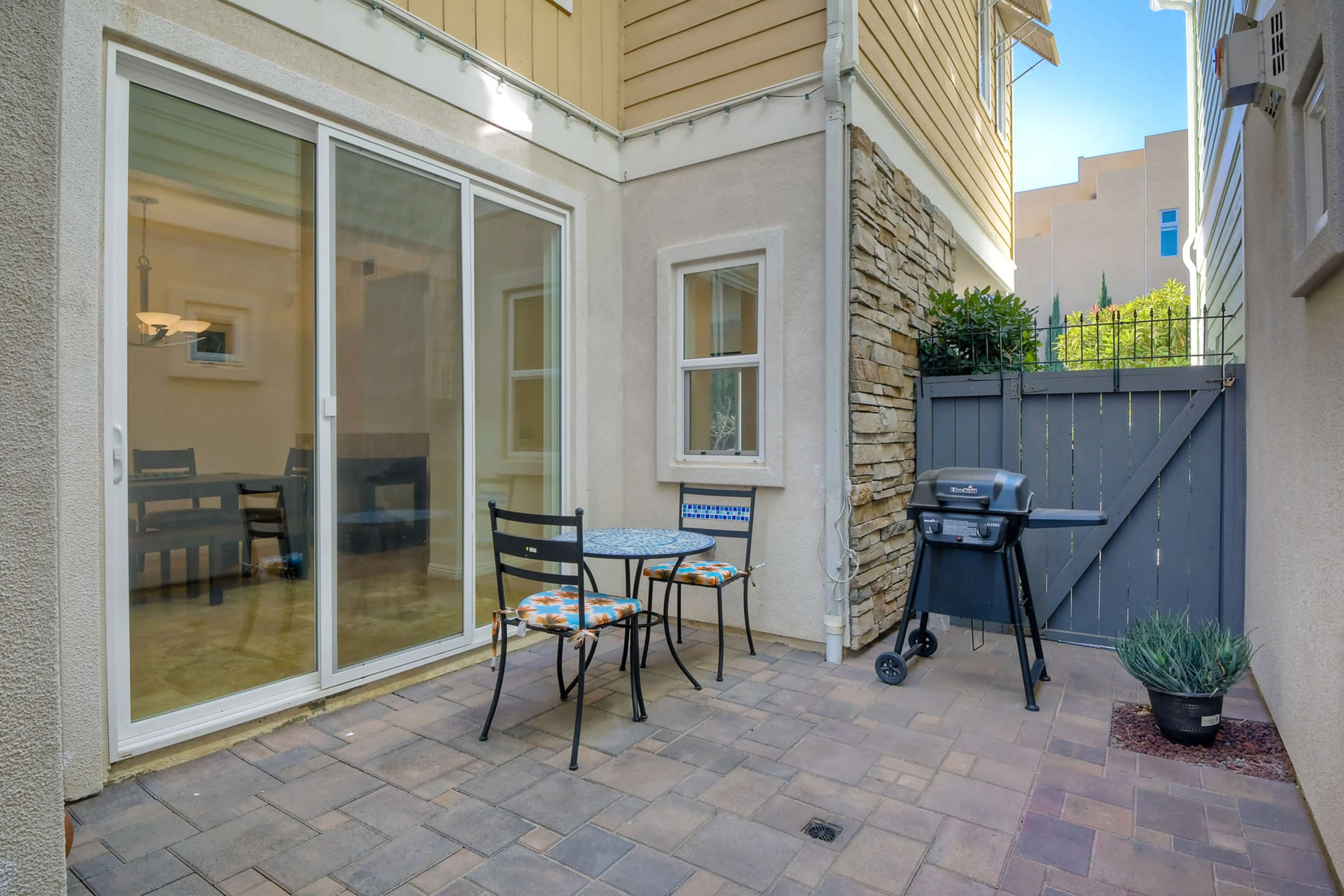 A small outdoor patio features a round table with two chairs and a grill next to a stone wall and sliding glass doors leading into a house.