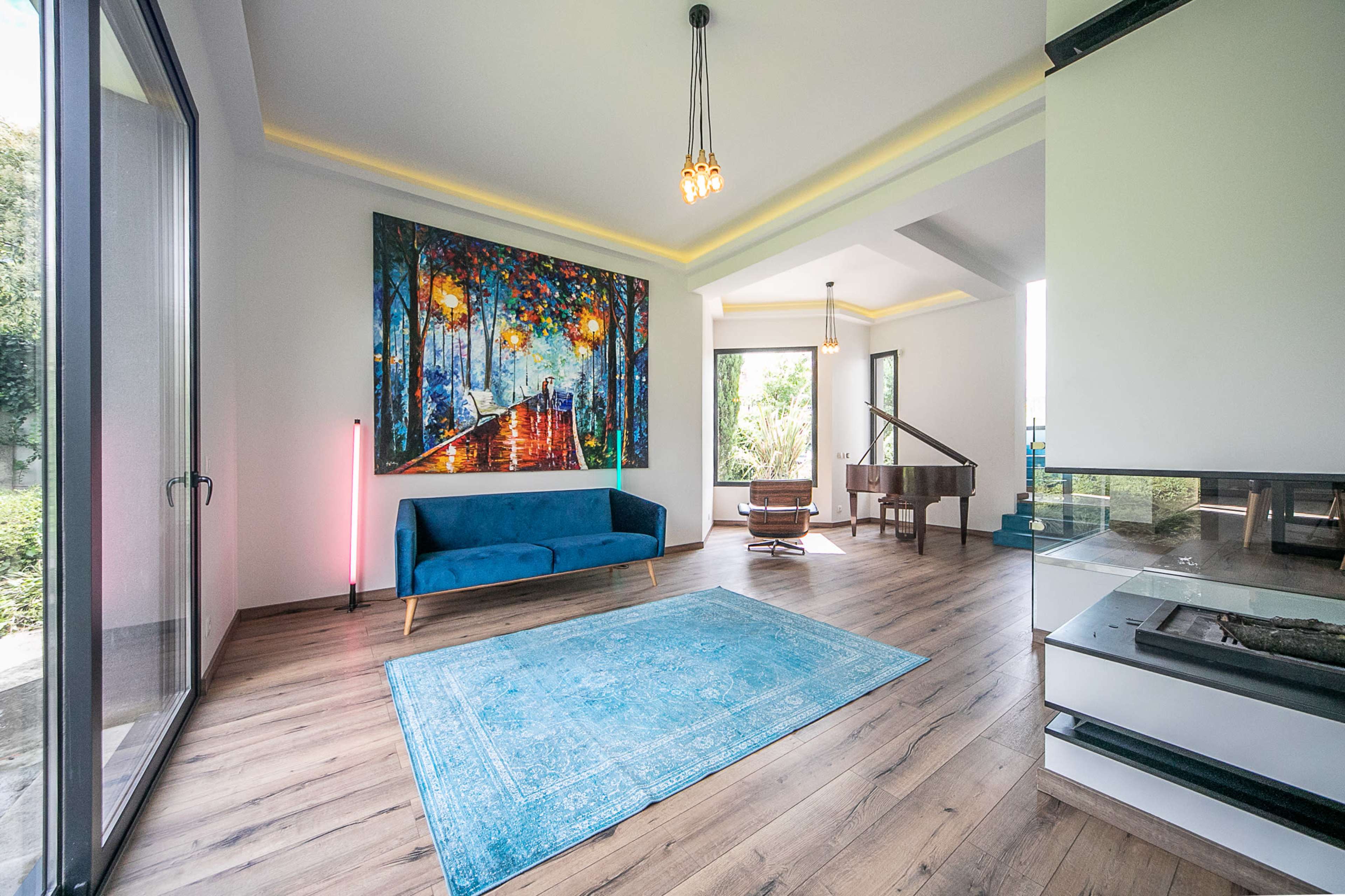 A living room features a blue couch, a colorful painting on the wall, a grand piano, and large windows that let in natural light.