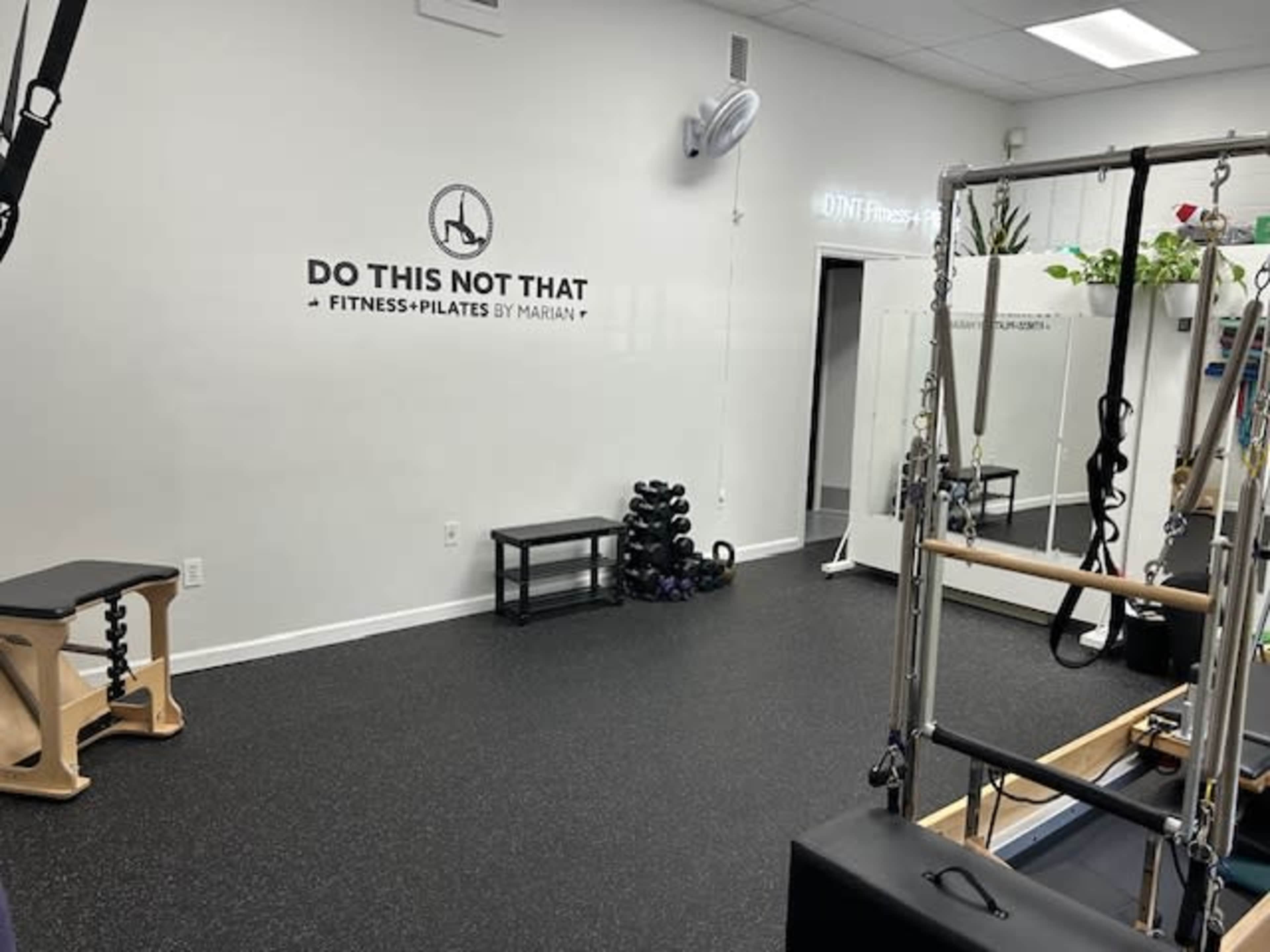 A fitness studio equipped with Pilates apparatus, including a reformer and free weights, with a sign on the wall that reads "DO THIS NOT THAT FITNESS + PILATES BY MARIAN."