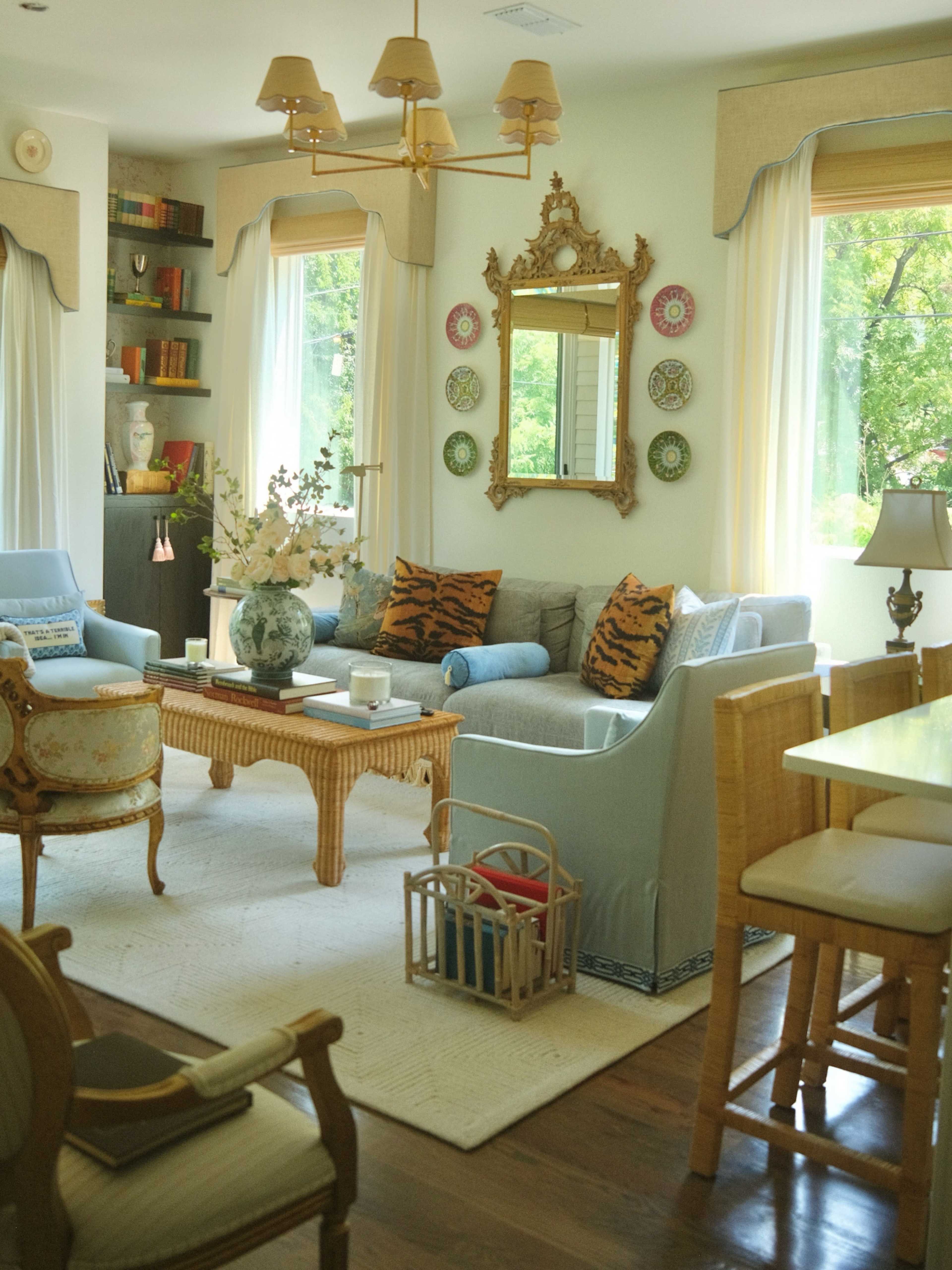 The image shows a well-decorated living room featuring a mix of seating arrangements, a coffee table with a floral centerpiece, and large windows letting in natural light.