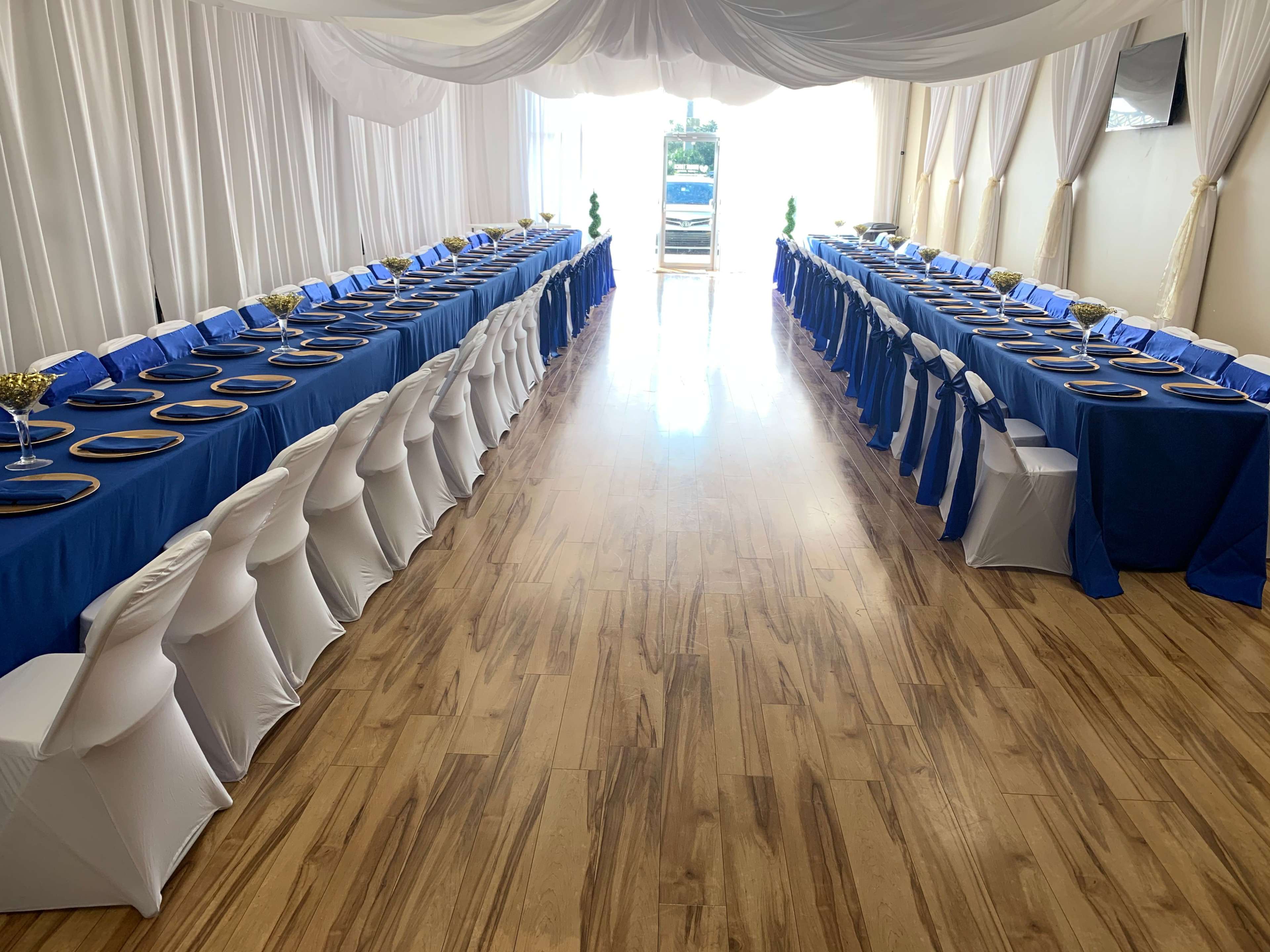 A spacious dining area features two long tables draped in blue and white linens, arranged with gold centerpieces and white chairs on polished wooden flooring.