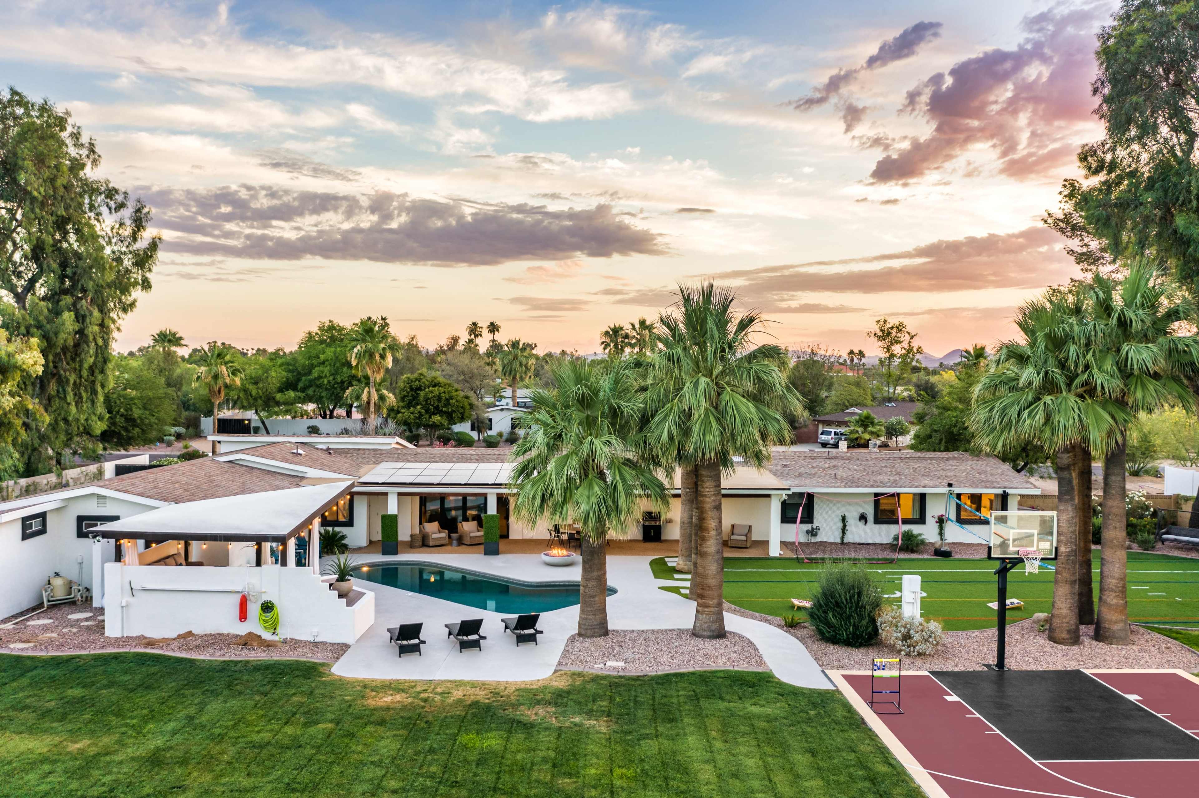 The image shows a spacious modern home with a swimming pool, surrounded by palm trees and a basketball court in a lush green yard at sunset.