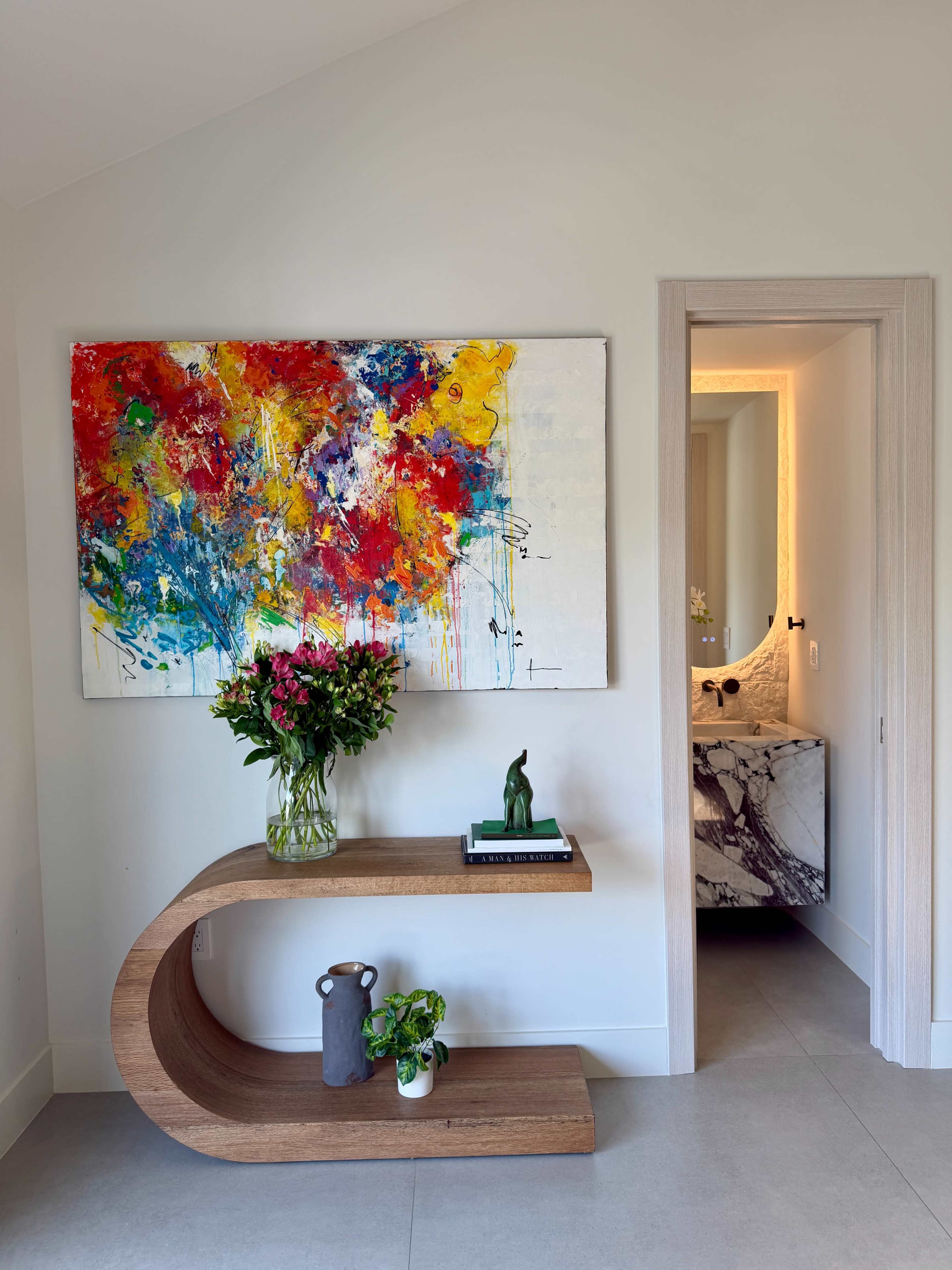 The image shows a modern entryway featuring a curved wooden table adorned with a vase of flowers, a small plant, and a decorative item, along with a colorful abstract painting on the wall and a bathroom doorway in the background.