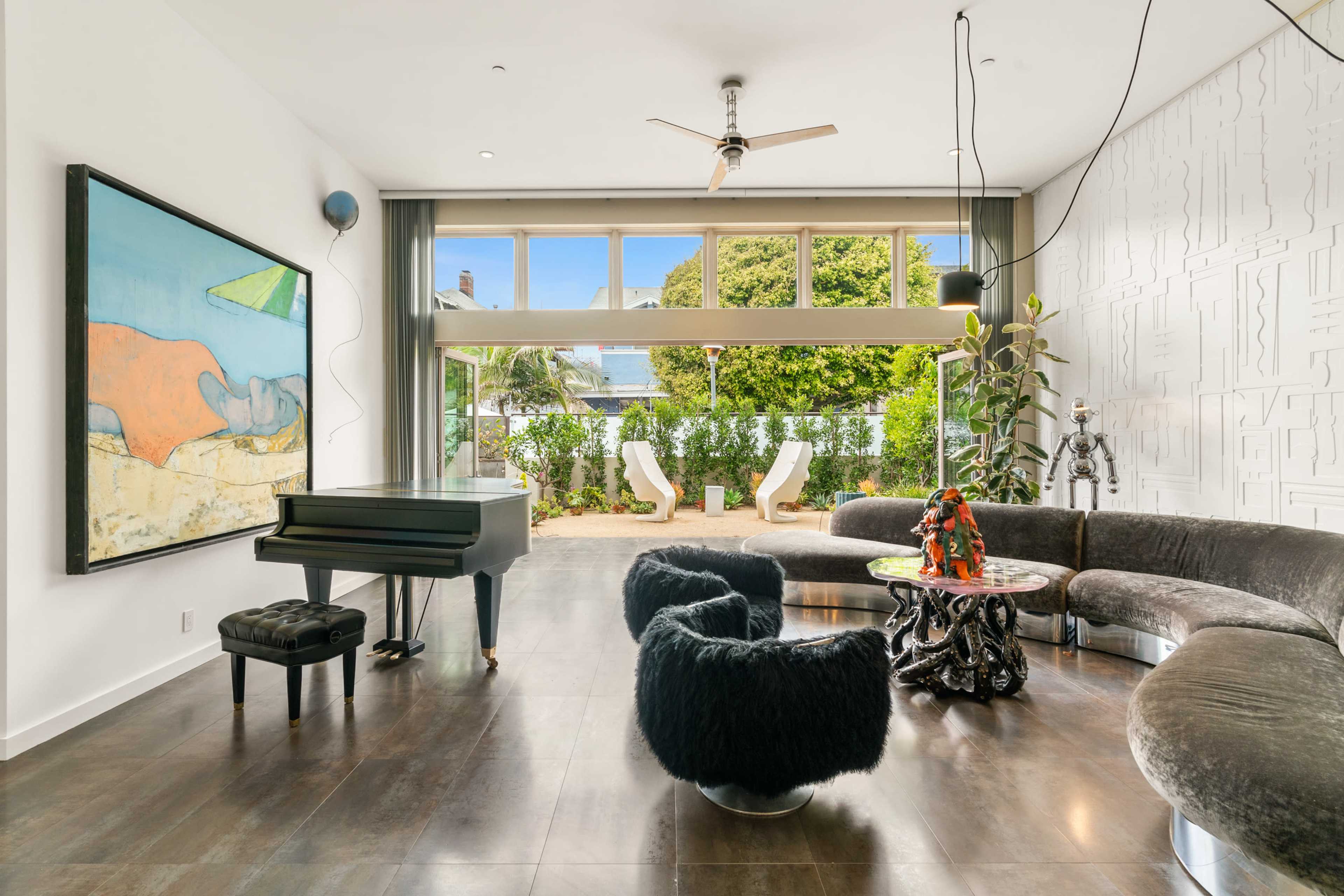 A modern living room featuring a grand piano, curved sofa, and large glass windows overlooking a garden.