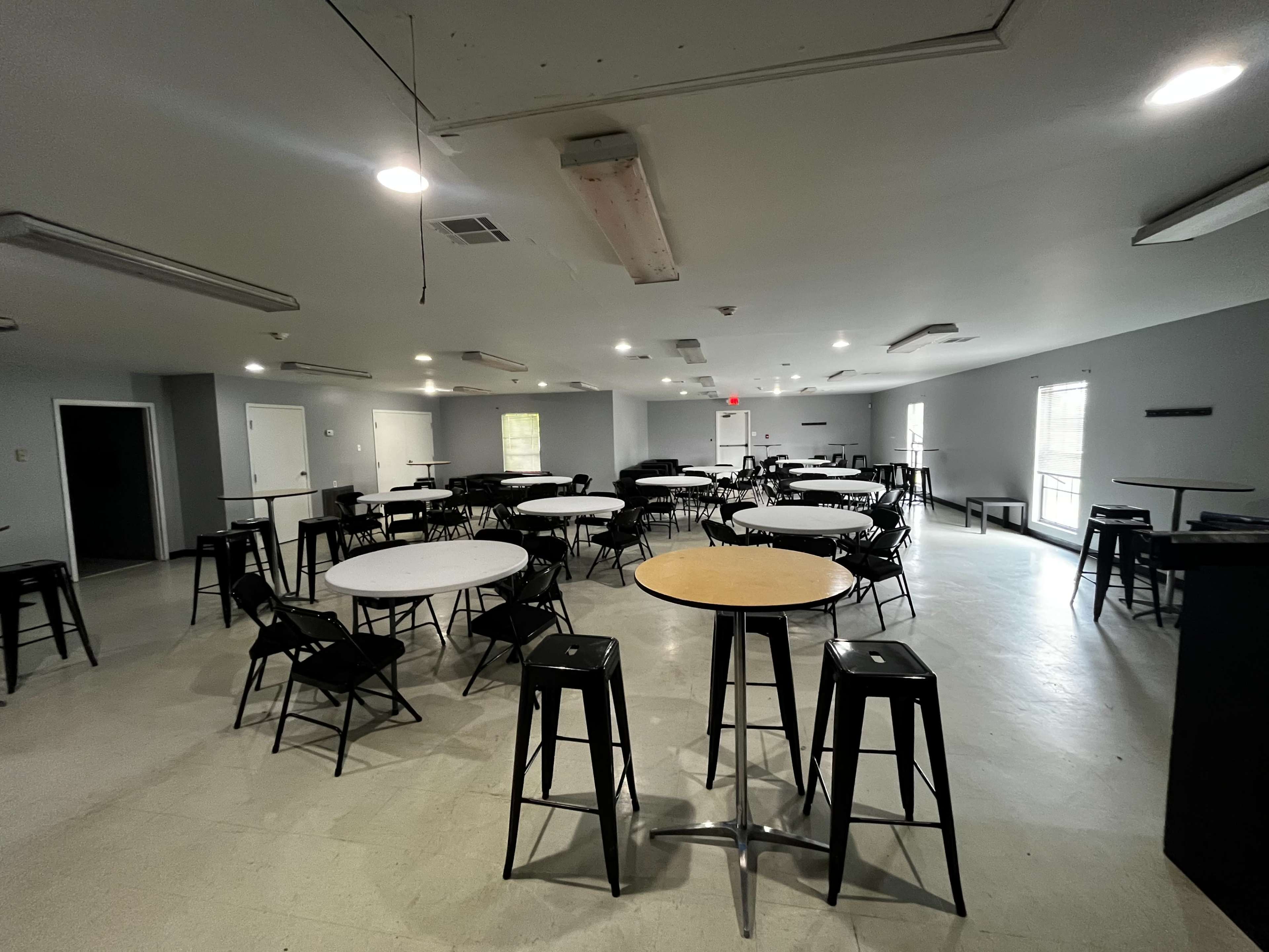 The image shows a spacious, well-lit room furnished with several round and rectangular tables surrounded by black chairs.