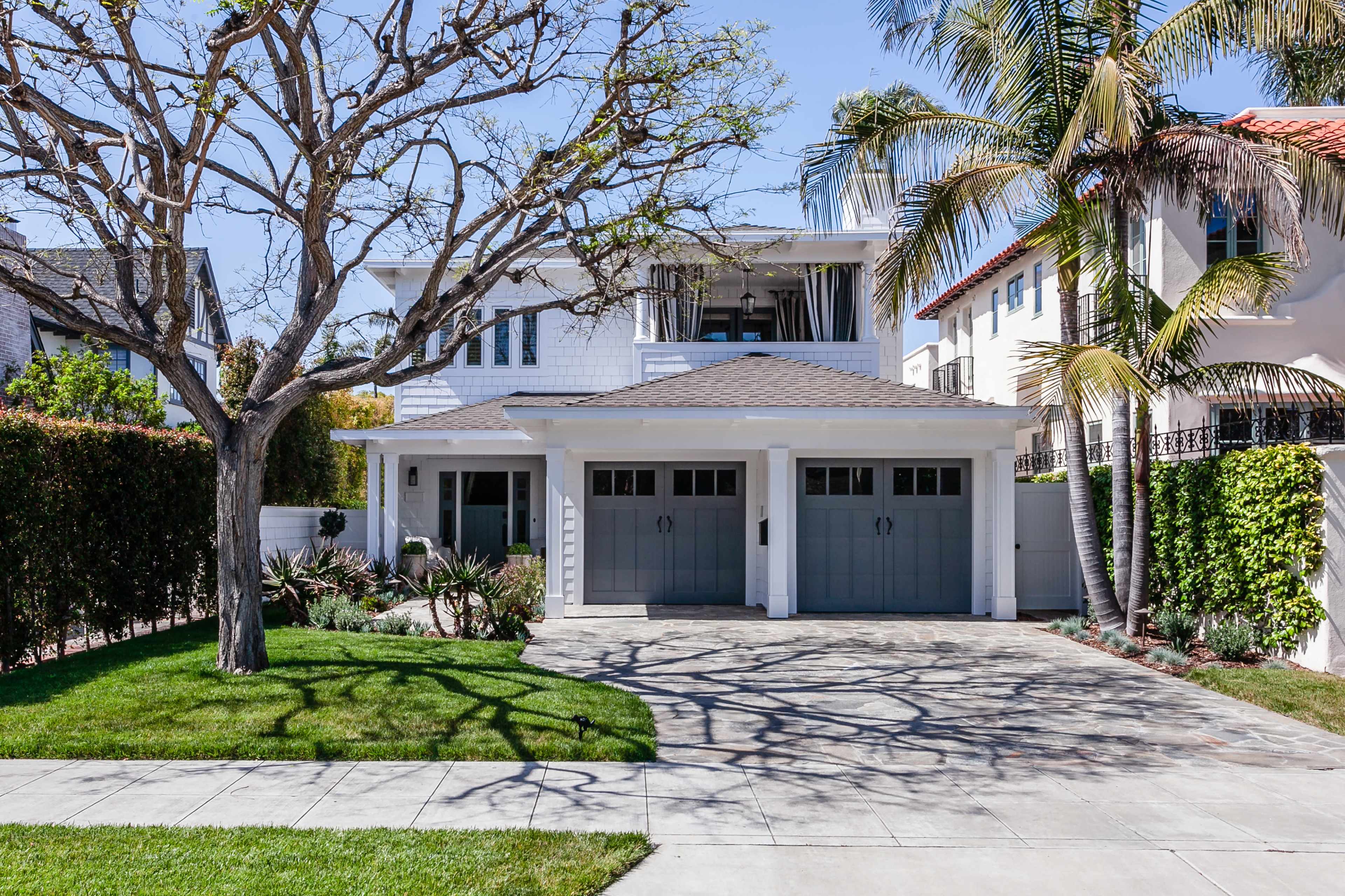 The image shows a two-car garage with gray doors in front of a two-story white house, surrounded by trees and manicured landscaping.