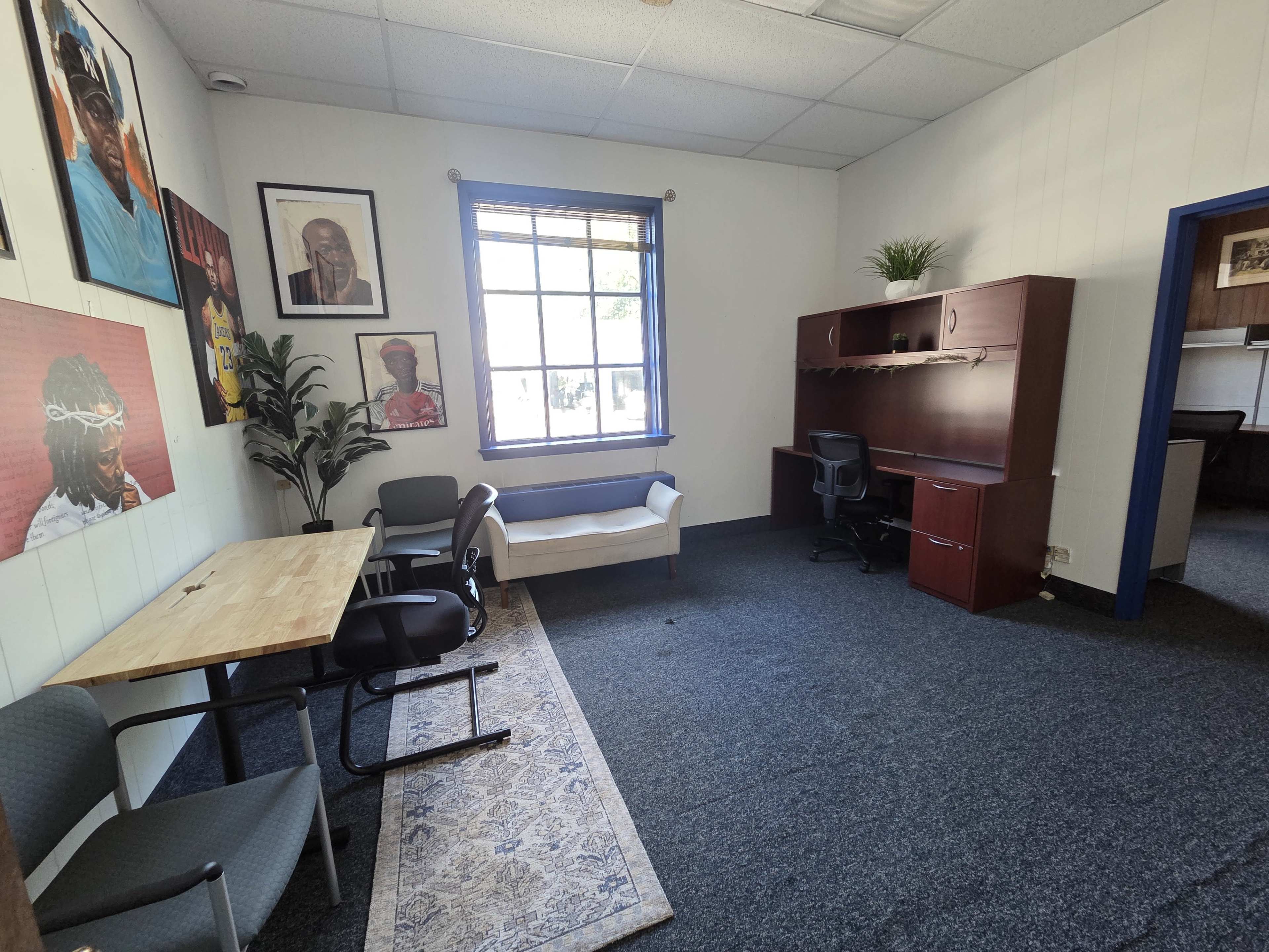 The image shows a small office space with a wooden table, two chairs, a white couch, a desk with a chair, and framed artwork on the walls.