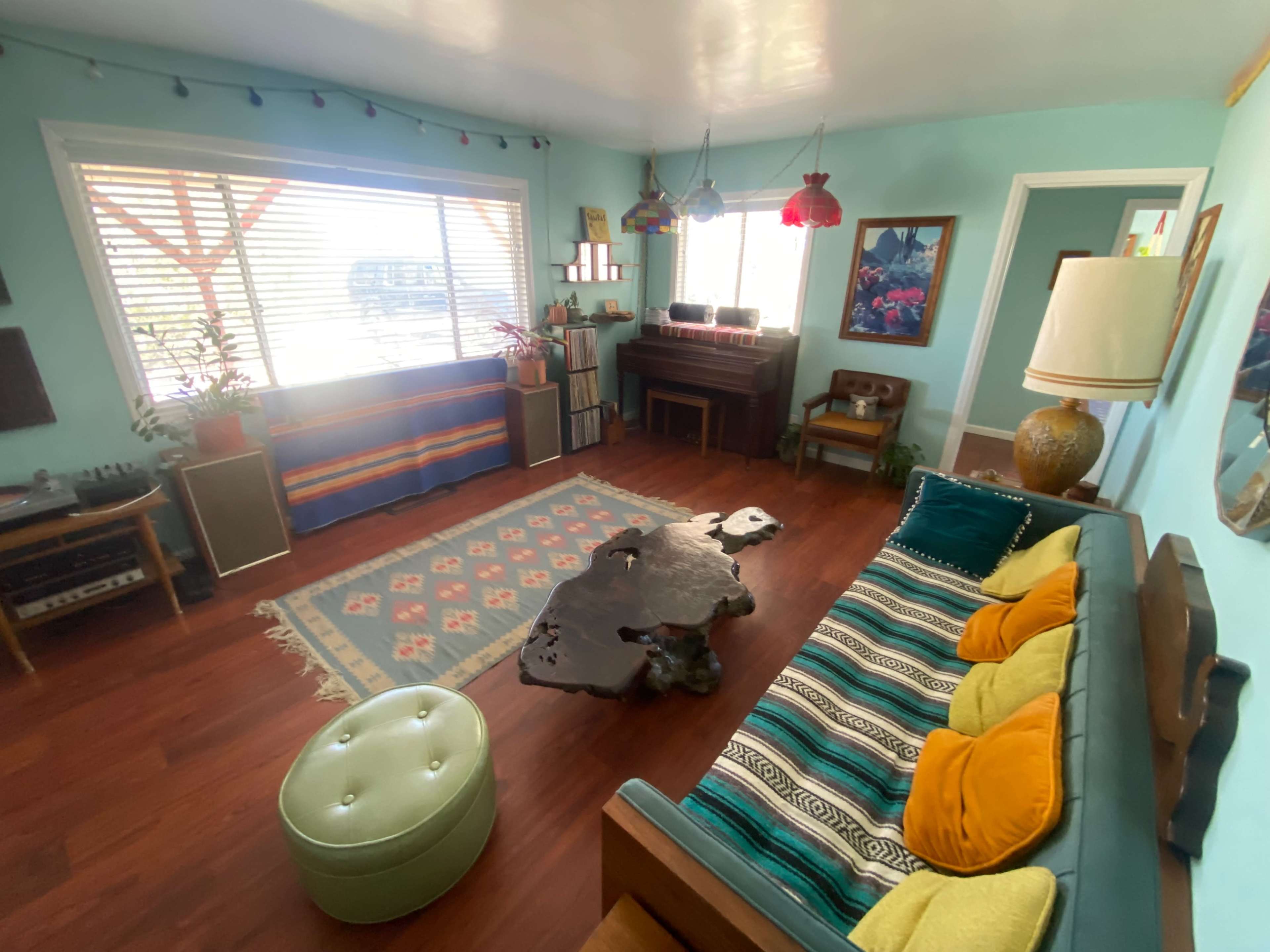 A brightly colored living room features a wooden sofa, a green ottoman, a coffee table with a unique shape, and various decorative elements, including plants and artwork, under large windows.