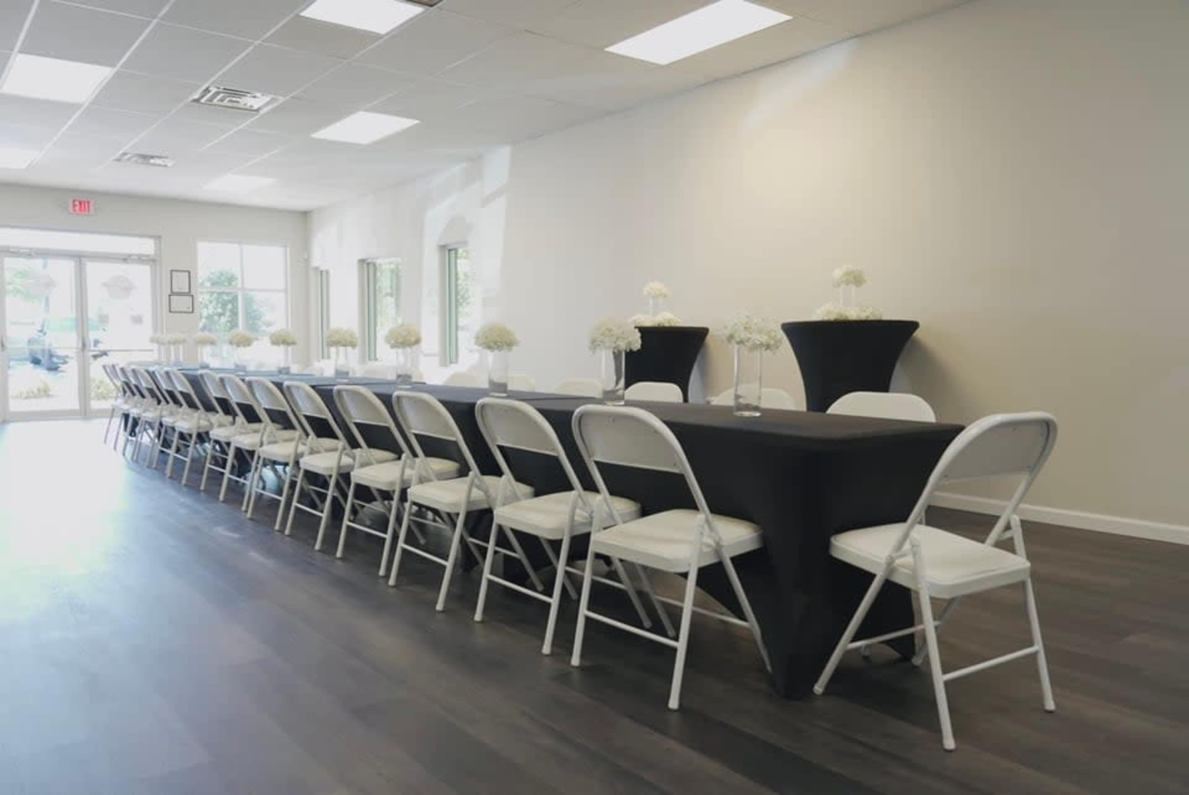 A long banquet table with white chairs is set up in a bright, empty room, decorated with small floral arrangements.