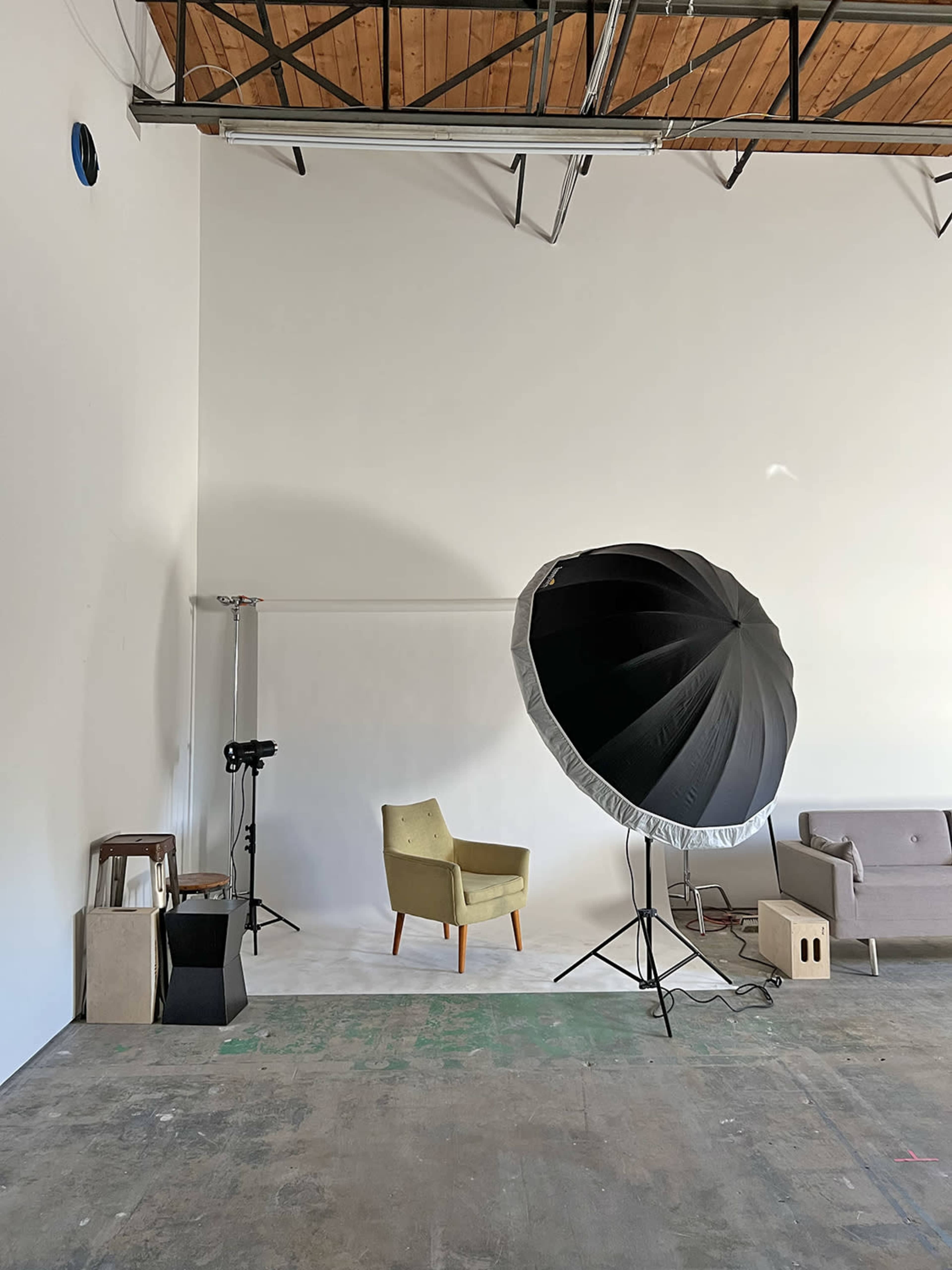 A minimalist studio space featuring a green upholstered chair, a gray couch, a large softbox light, and various props arranged against a white wall.