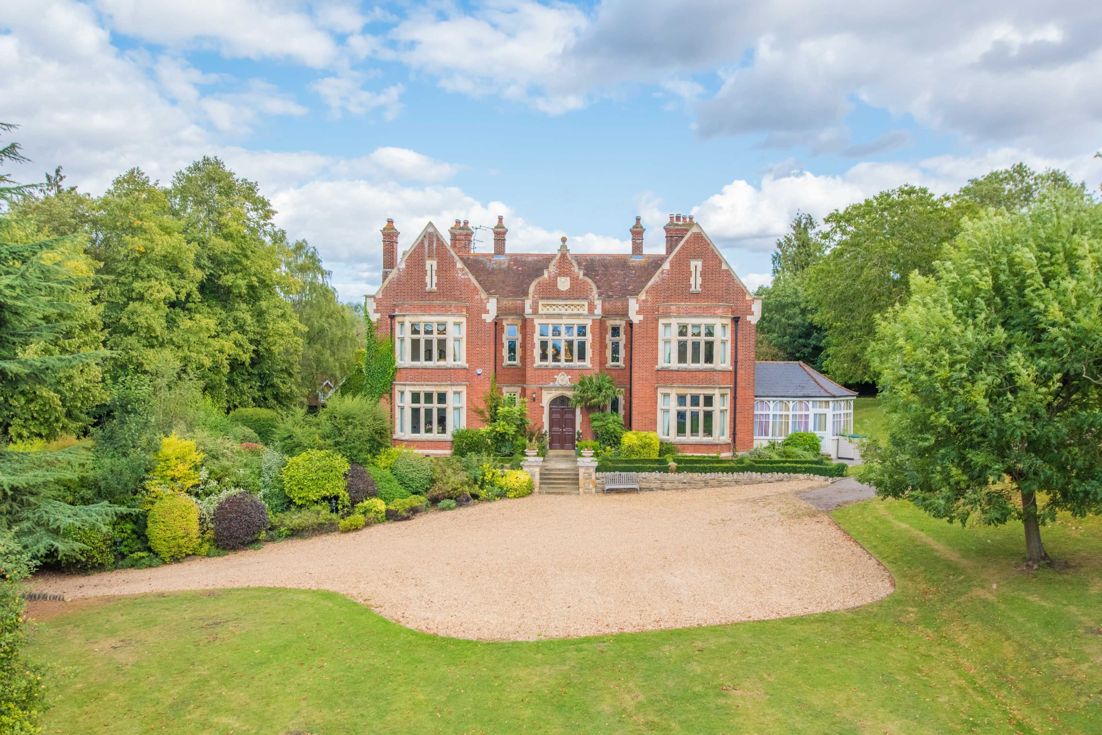 A large red brick mansion is surrounded by well-maintained gardens and trees, set against a partly cloudy sky.