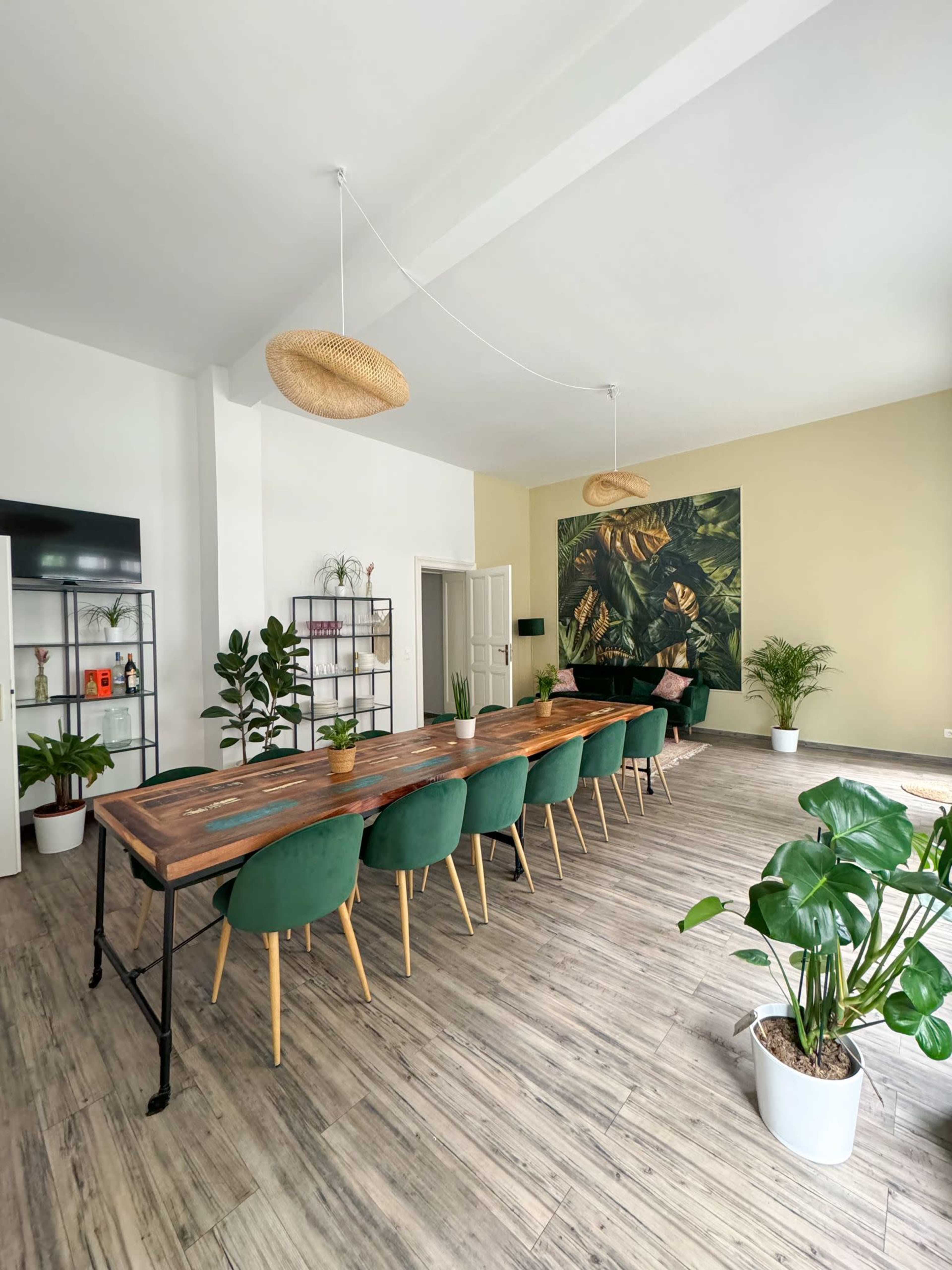 A spacious dining area features a long wooden table surrounded by green chairs, potted plants, and decorative wall art with tropical leaves.