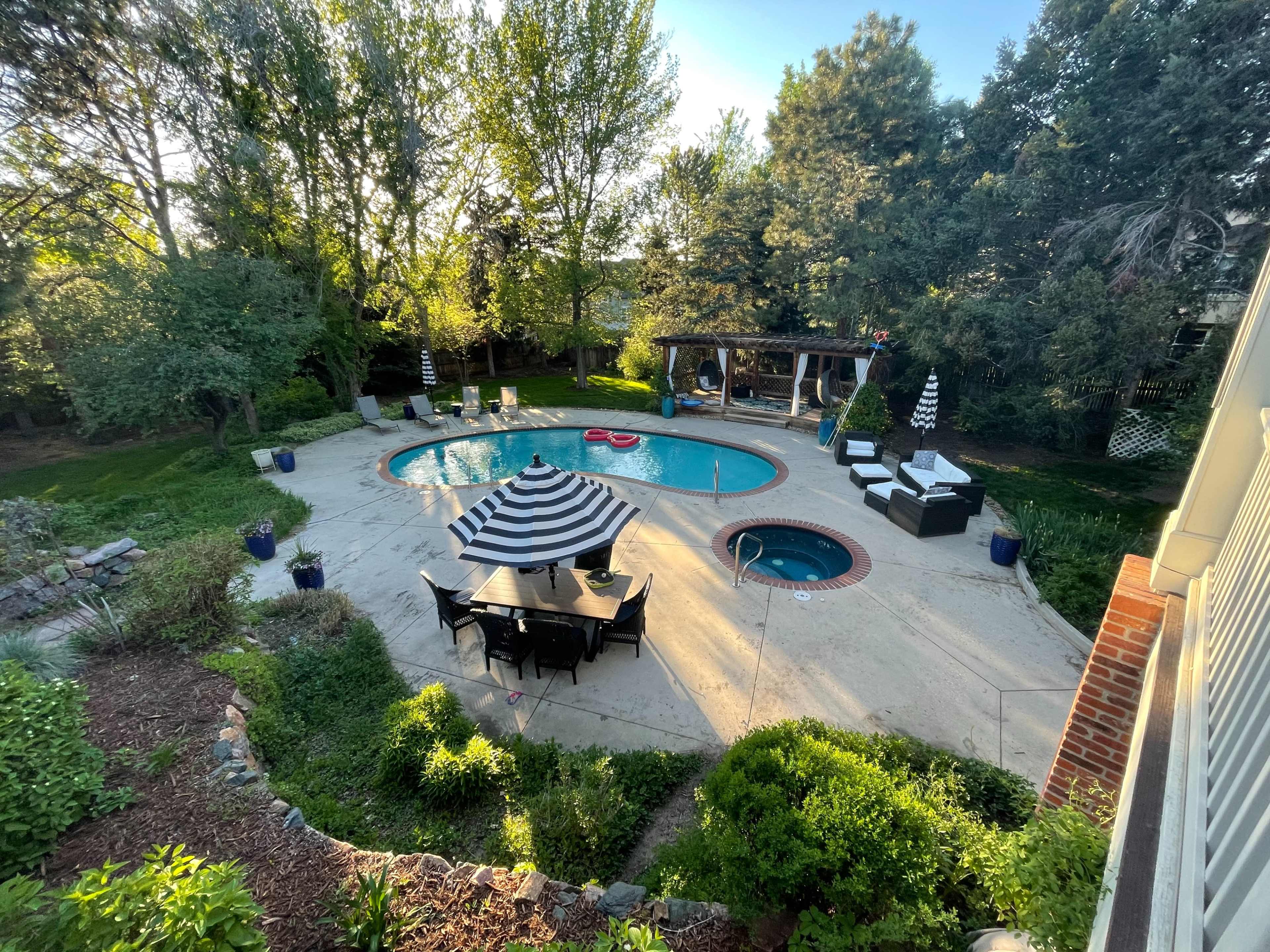 The image shows a backyard pool area with a swimming pool, a hot tub, striped umbrellas, and dining furniture surrounded by greenery.