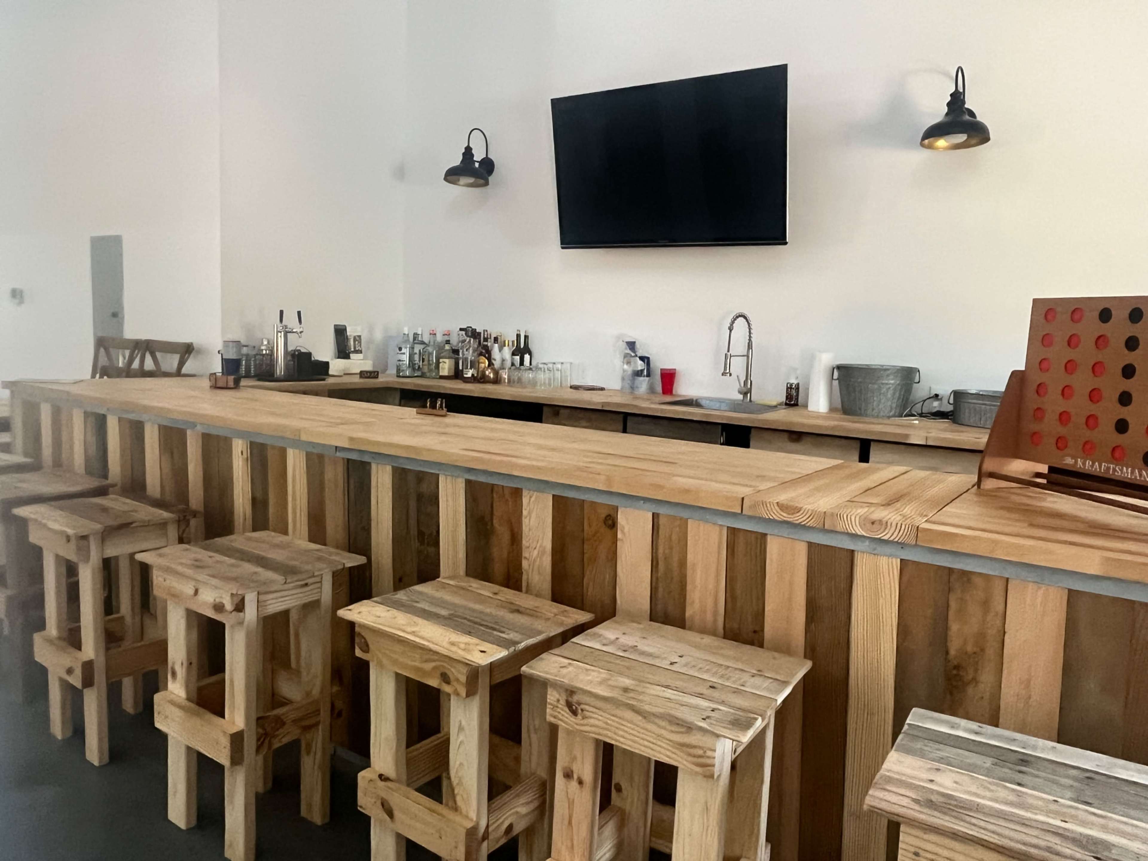 A wooden bar with stools is set against a plain wall, featuring a television and an assortment of bottles on the counter.