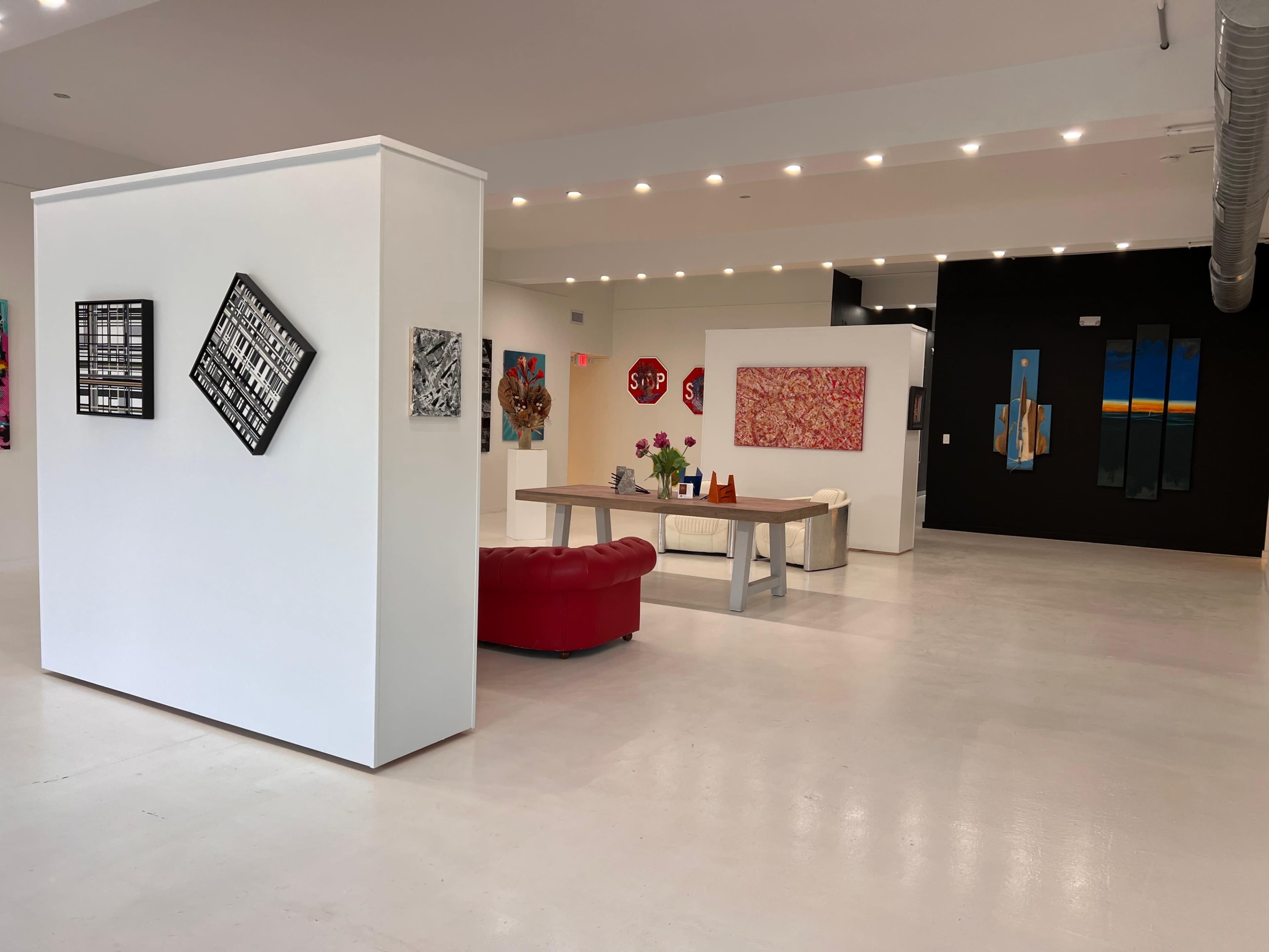 The image shows a modern art gallery with white walls, various artworks displayed, and a red couch in the foreground.