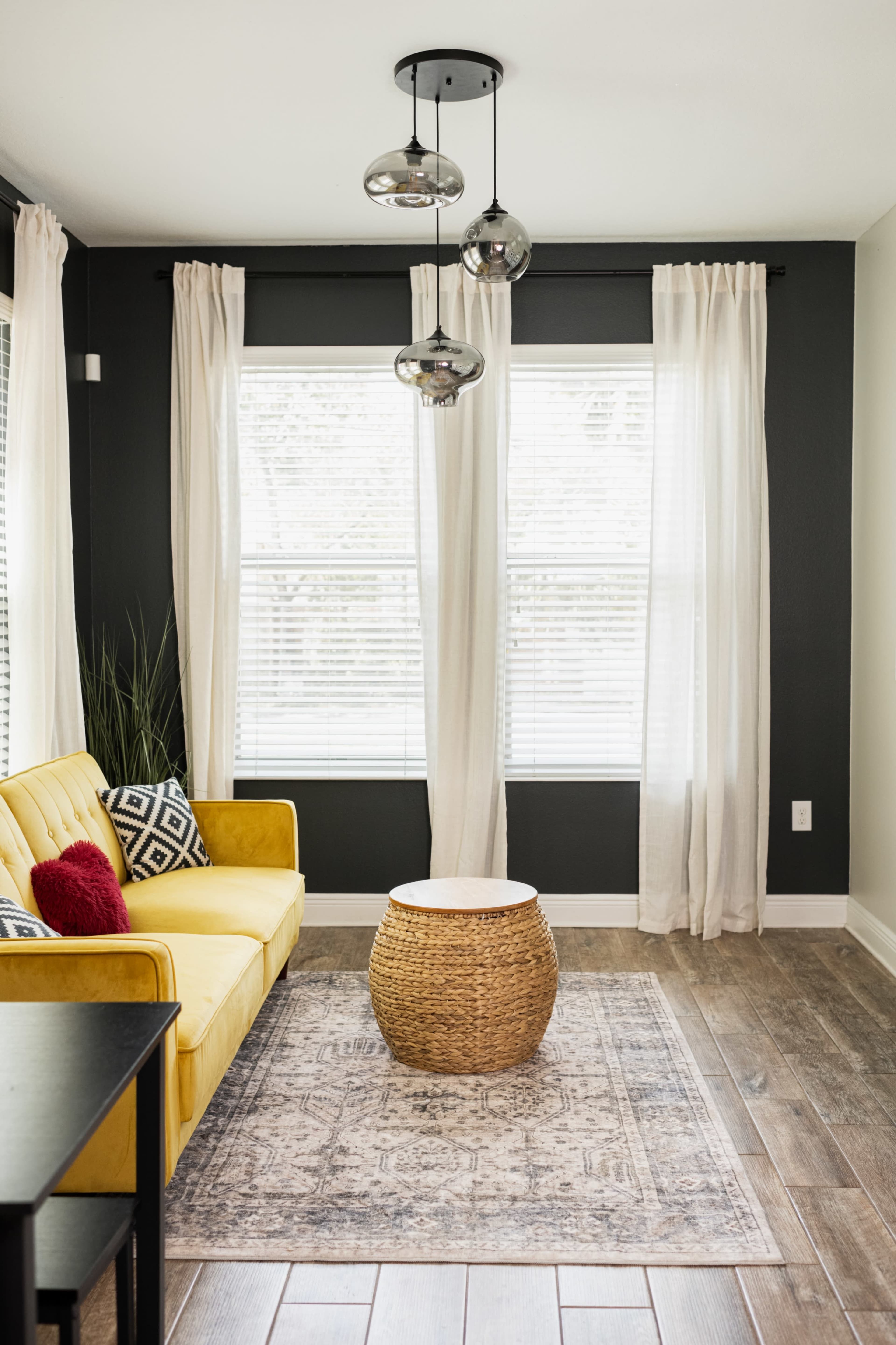 The image shows a cozy living room with a yellow sofa, a round woven coffee table, and large windows adorned with white curtains.