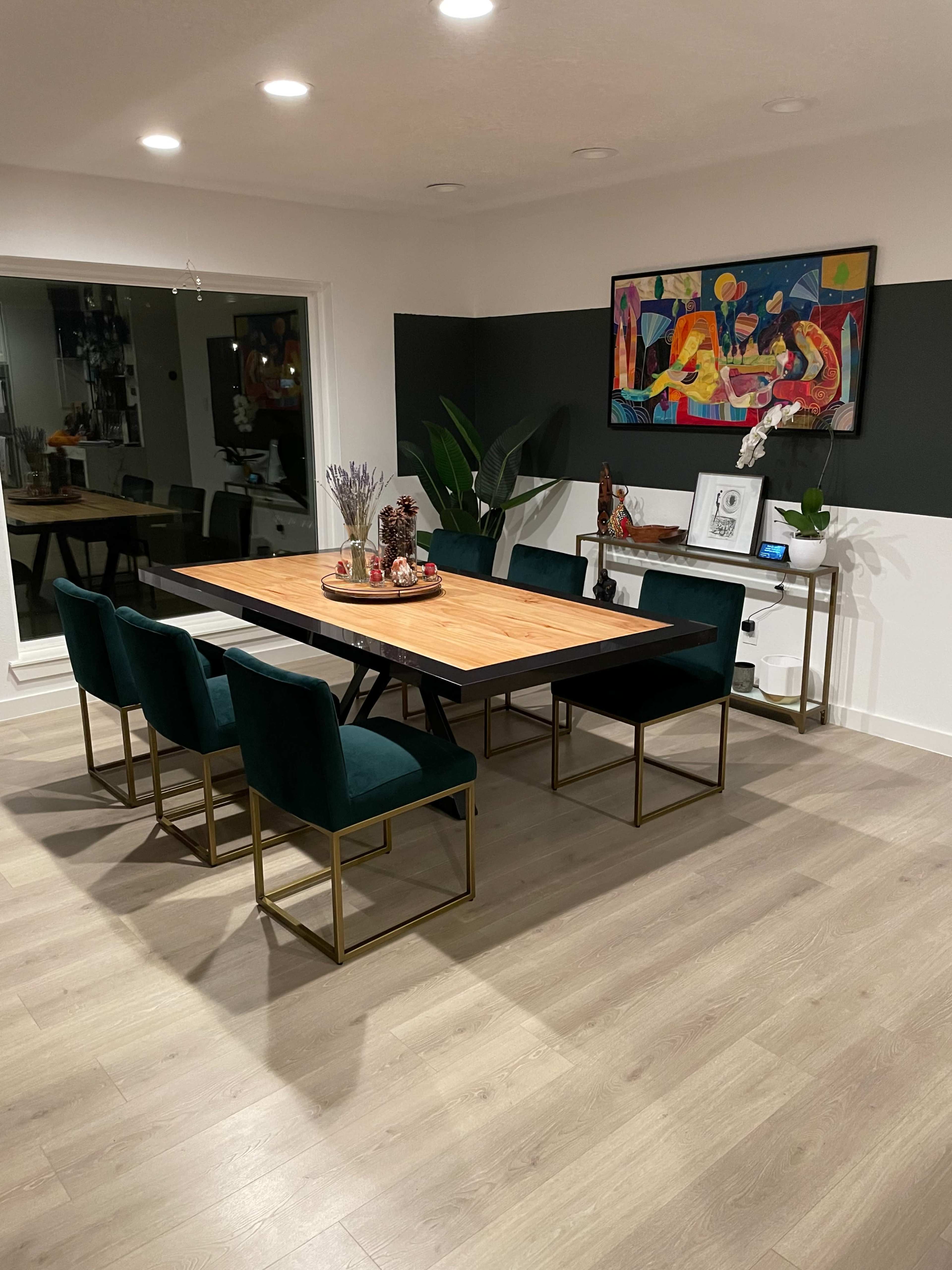A modern dining room features a wooden table surrounded by six green chairs, with a large artwork hanging on the wall and a console table displayed against a contrasting black backdrop.