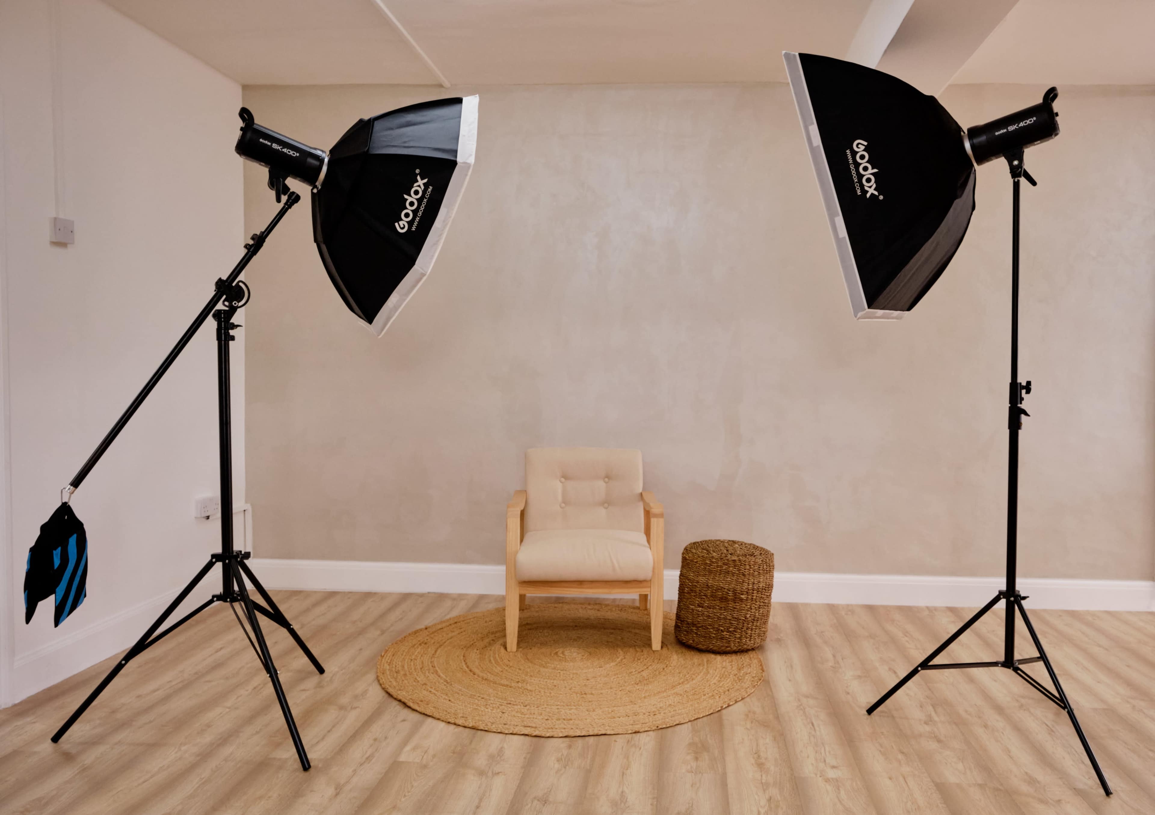 A minimalist photography studio with two softbox lights on stands facing a small, light-colored chair and a round woven rug.