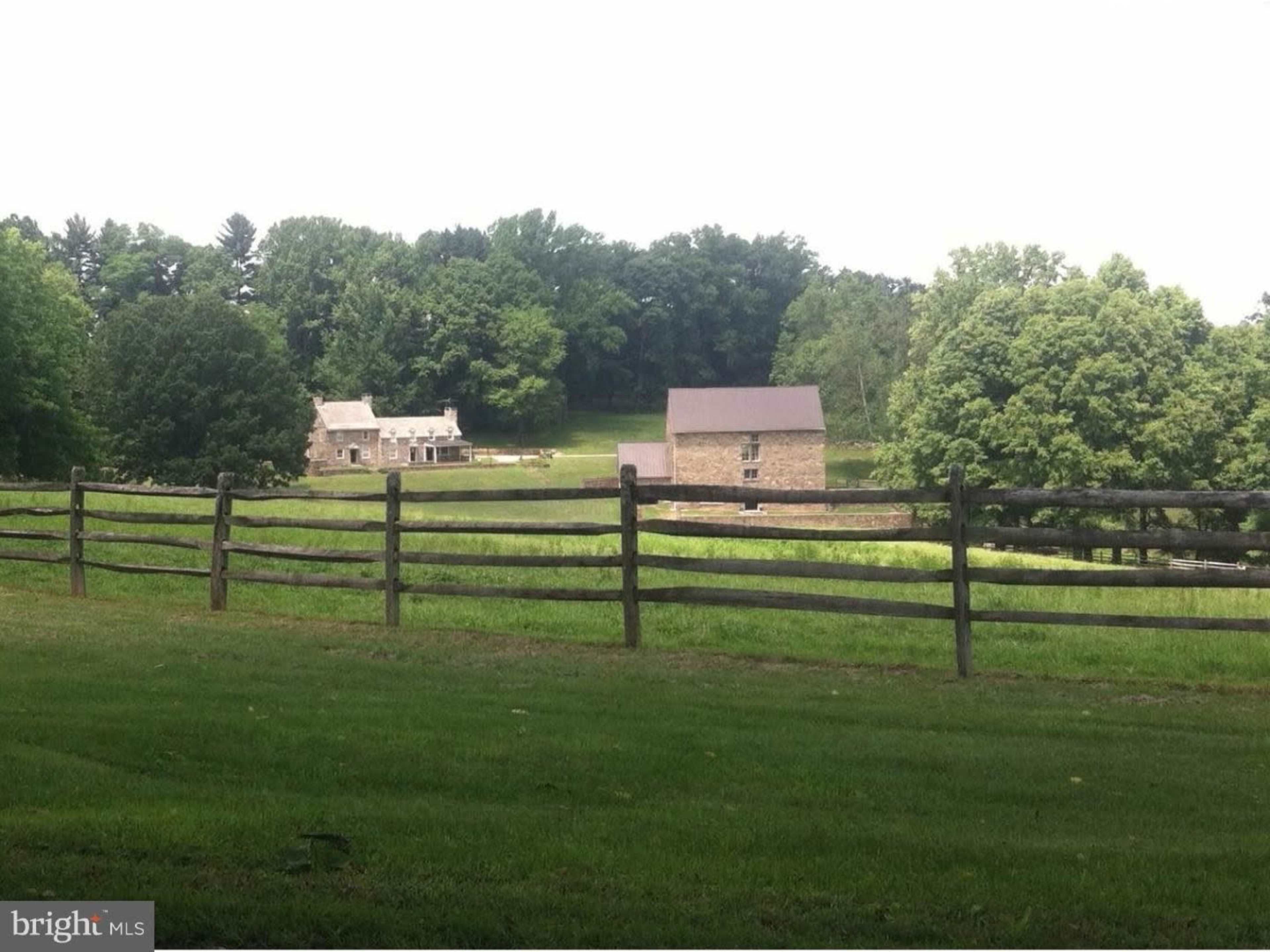 The image shows a rural landscape with a wooden fence in the foreground and two stone buildings set against a backdrop of trees and open fields.