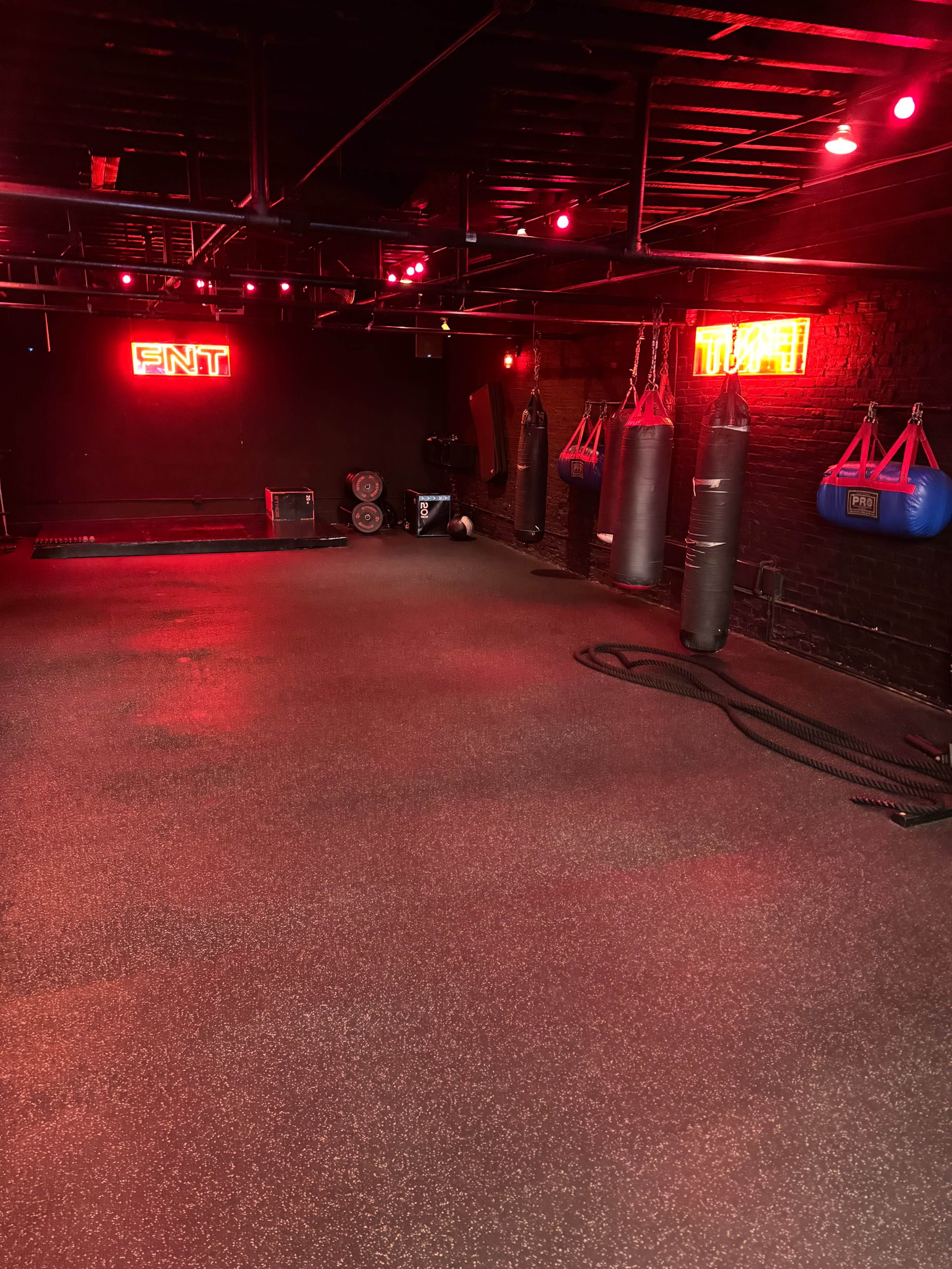 The image shows an empty gym space with red lighting, featuring boxing bags, exercise ropes, and neon signs on the walls.