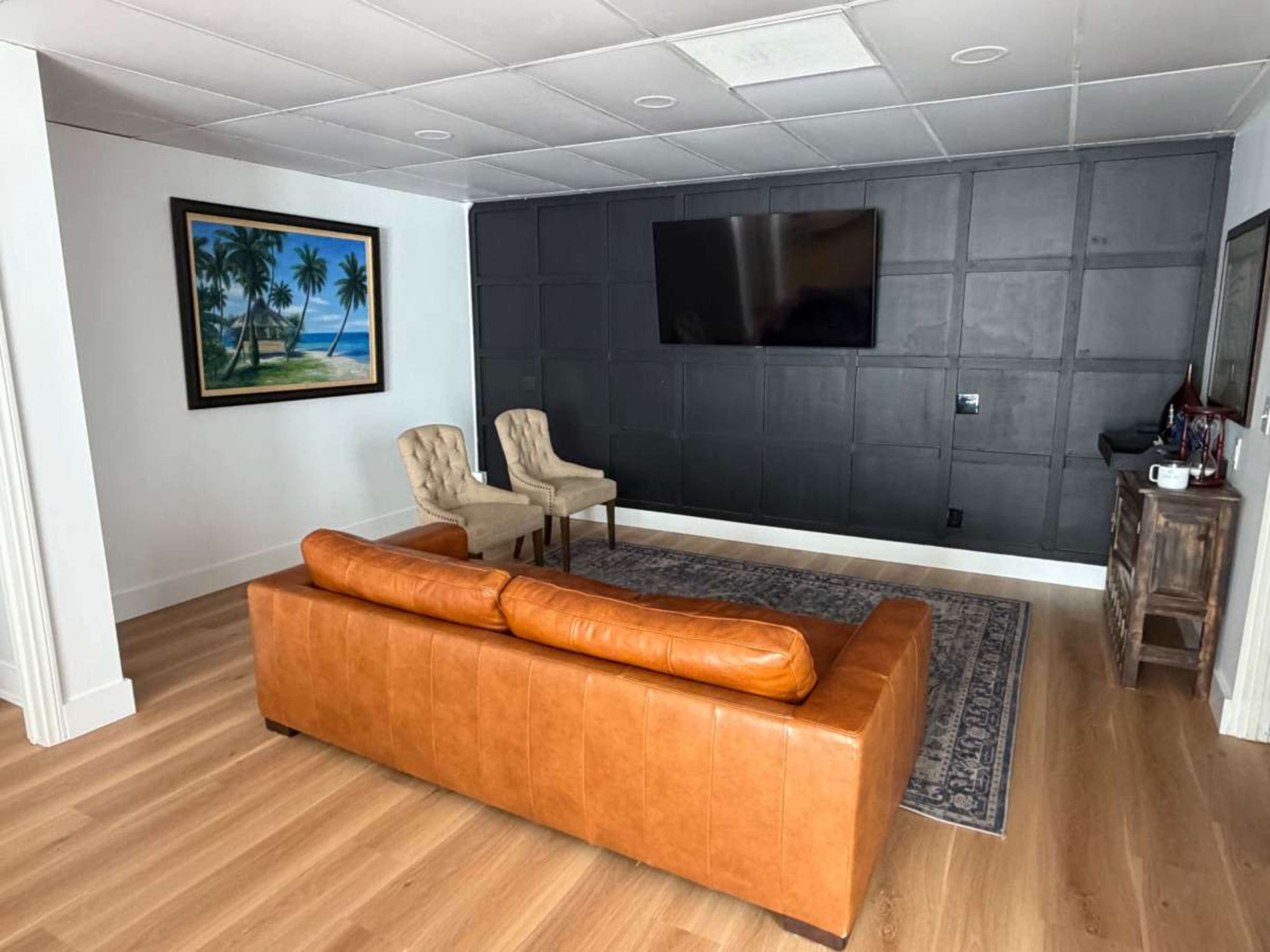 The image shows a modern living space with a brown leather sofa, two light-colored chairs, a large wall-mounted TV, and a framed painting of a tropical scene.