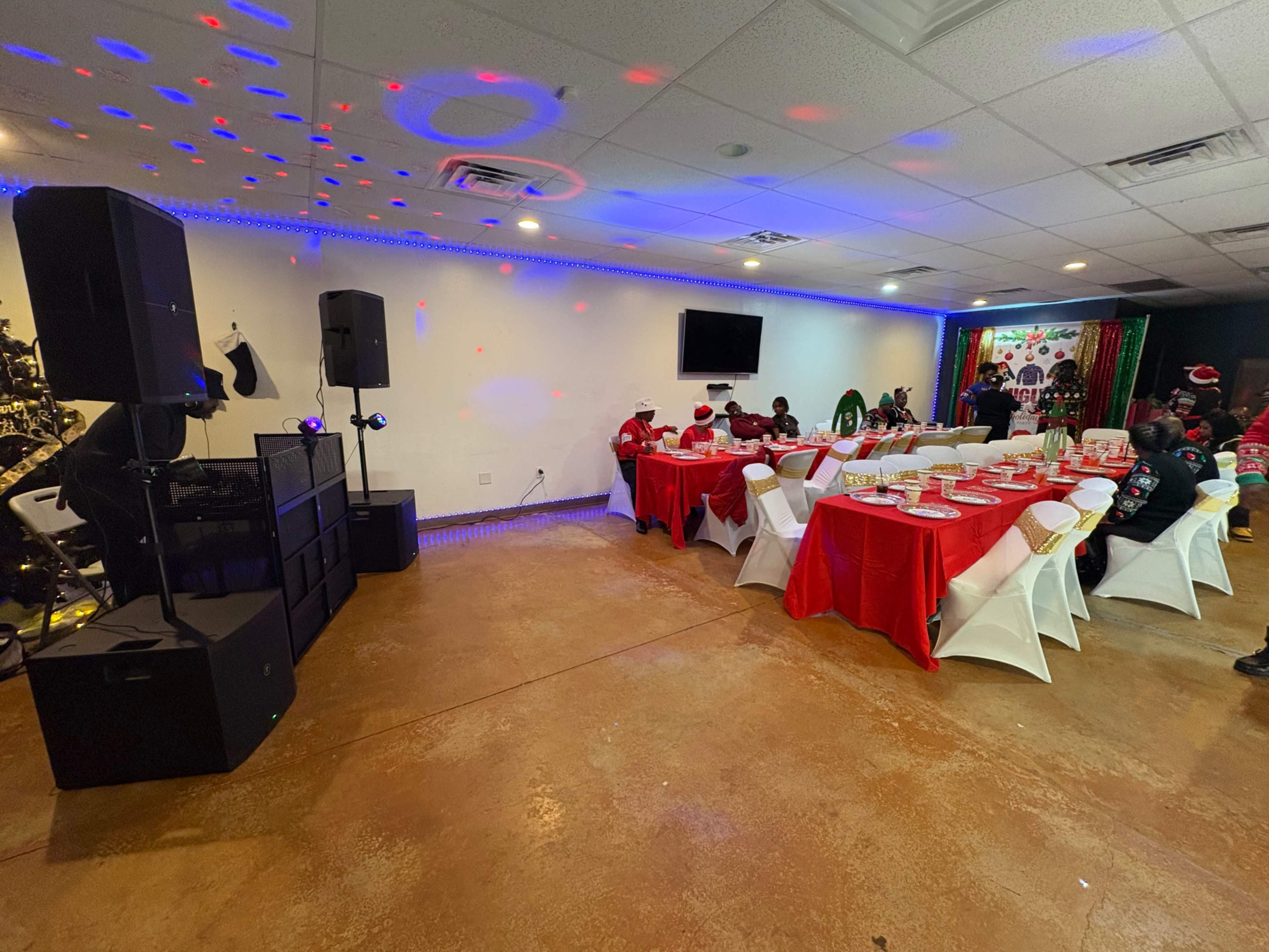 A decorated banquet room features a long table set with red tablecloths, surrounded by white chairs and festive lighting.