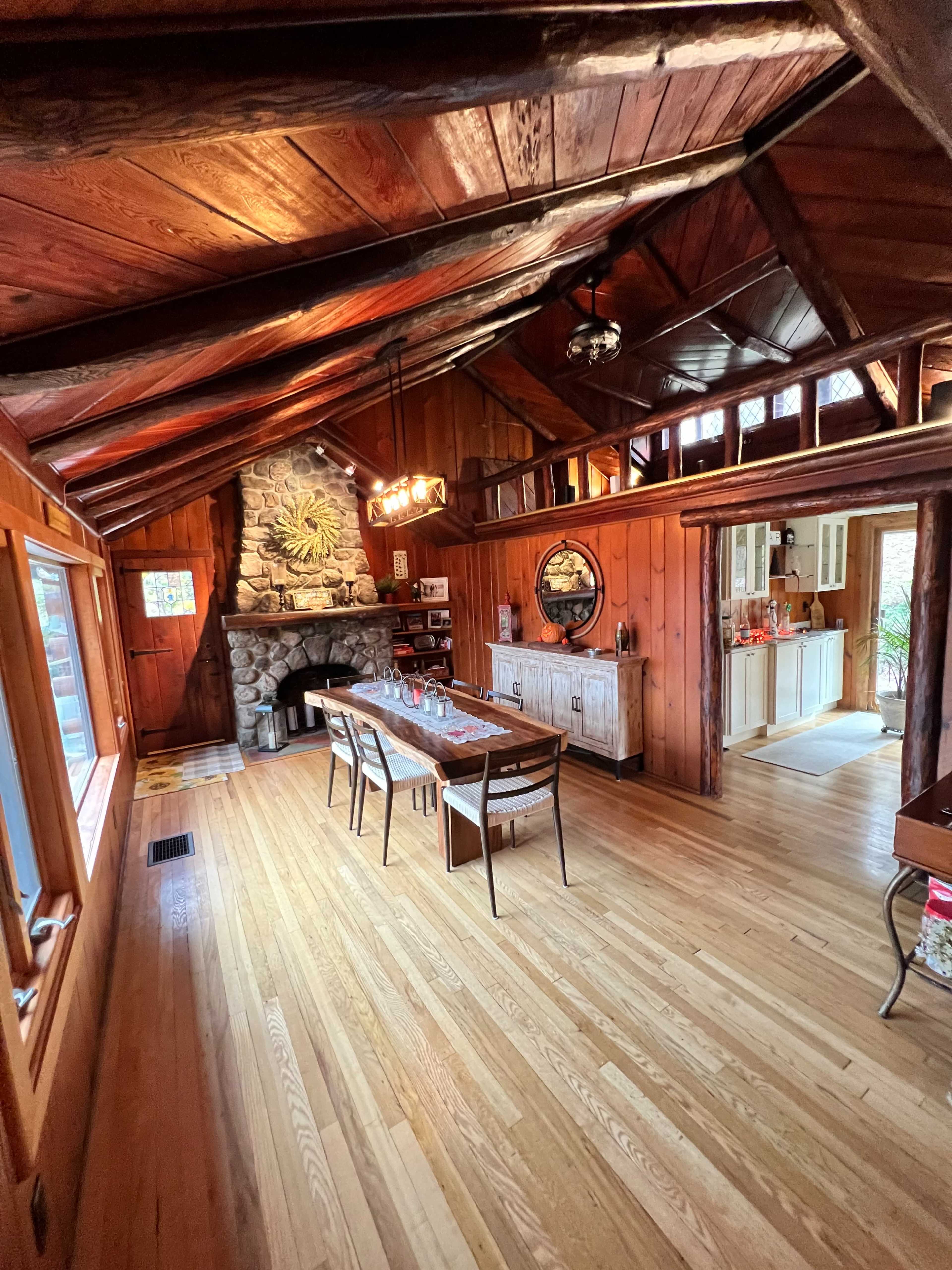 The interior of a wooden cabin features a dining area with a table, stone fireplace, and large windows.