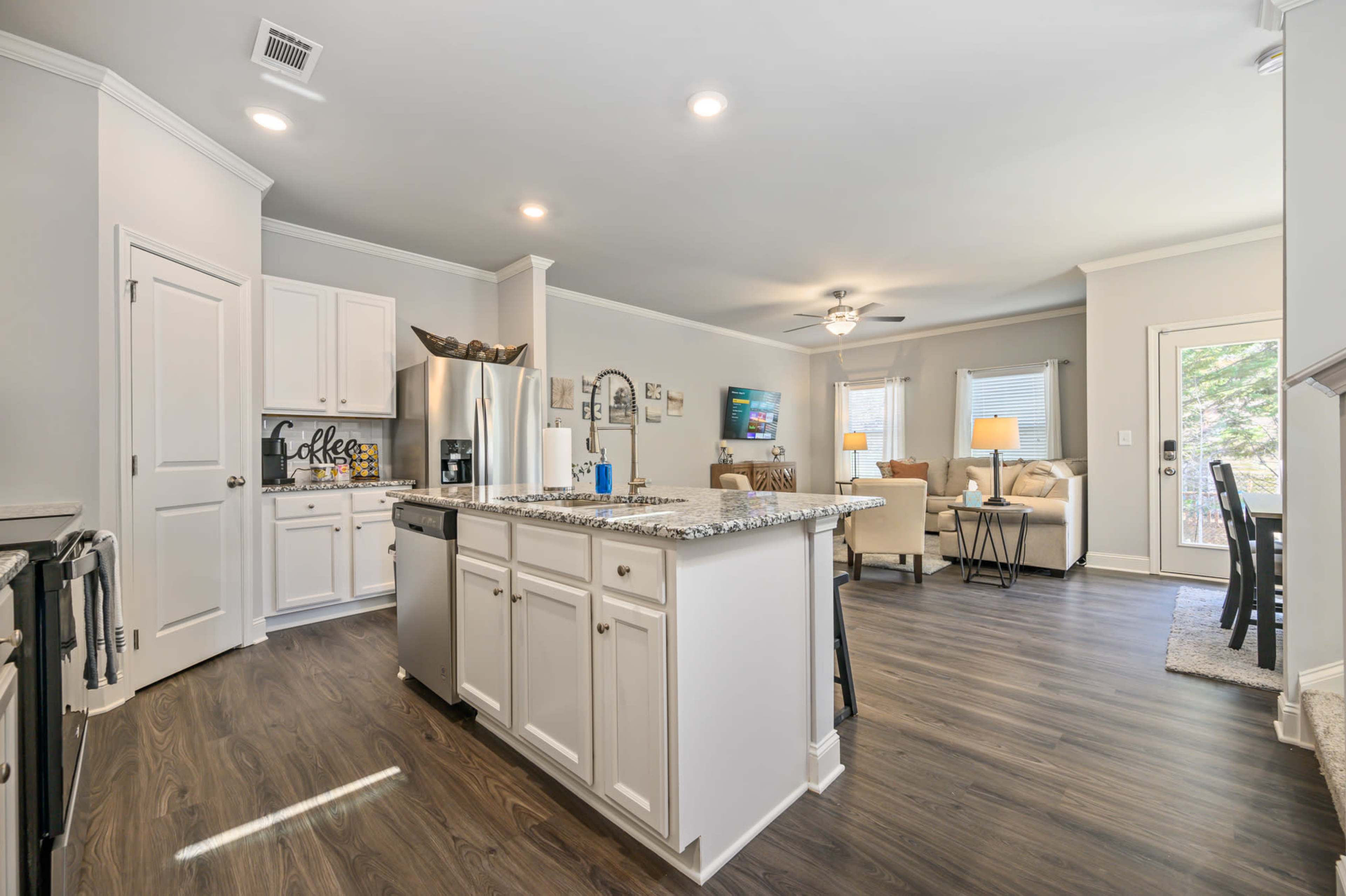 A modern kitchen with white cabinetry, a granite countertop island, stainless steel appliances, and an open layout leading to a living area.