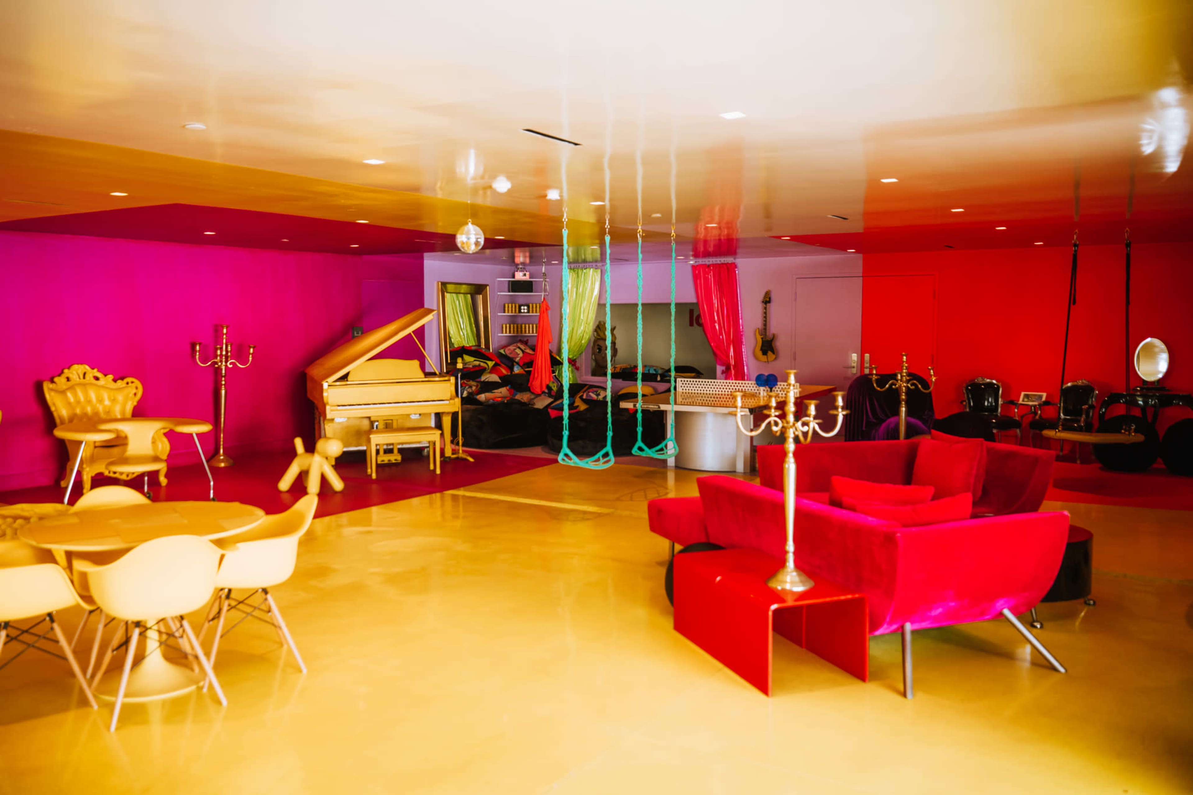 The brightly colored room features plush furniture, a grand piano, and decorative candelabras against a backdrop of vibrant pink and gold walls.