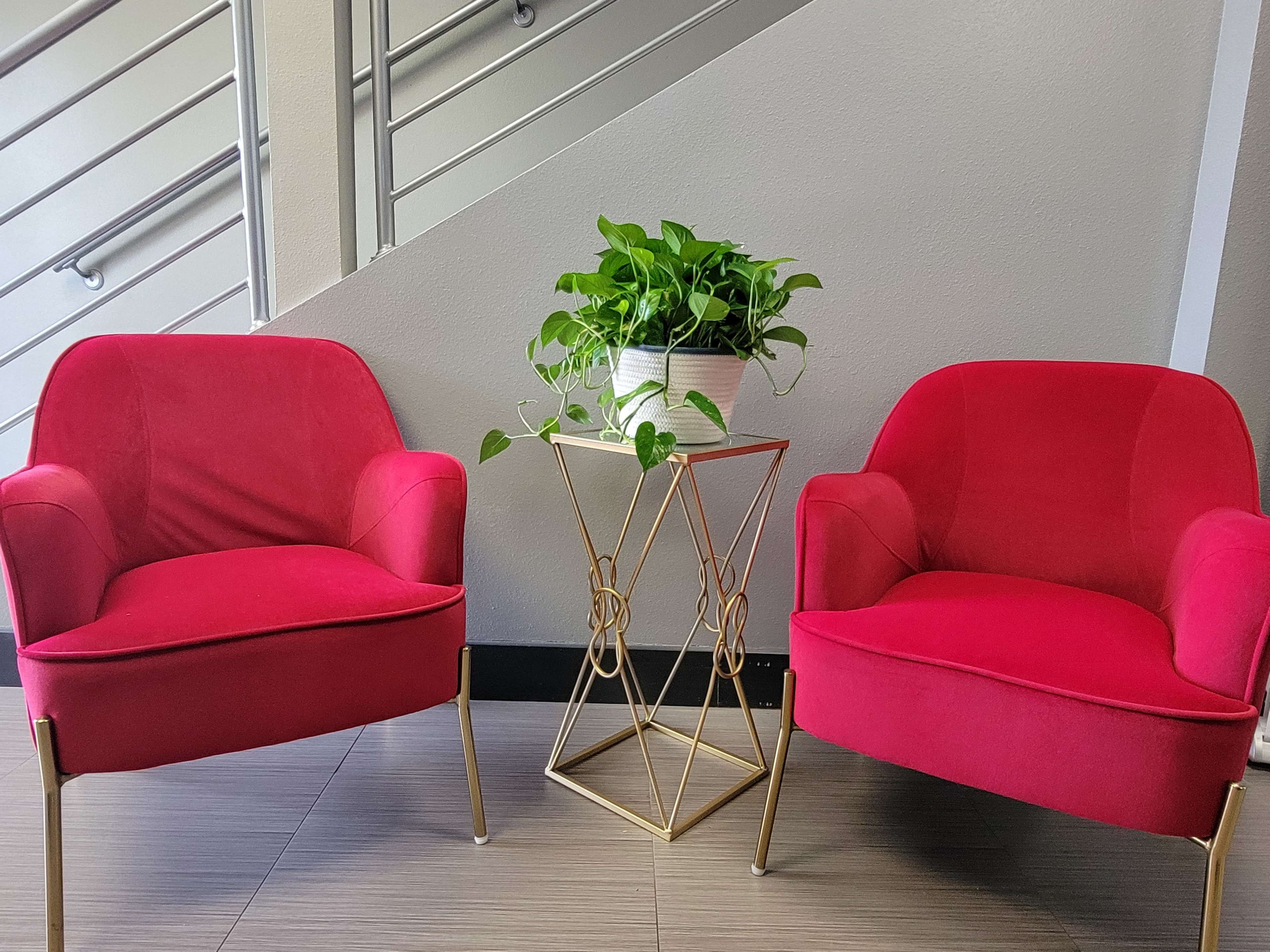 Two pink velvet chairs are positioned on either side of a gold metal side table, which holds a potted plant, against a wall near a staircase.
