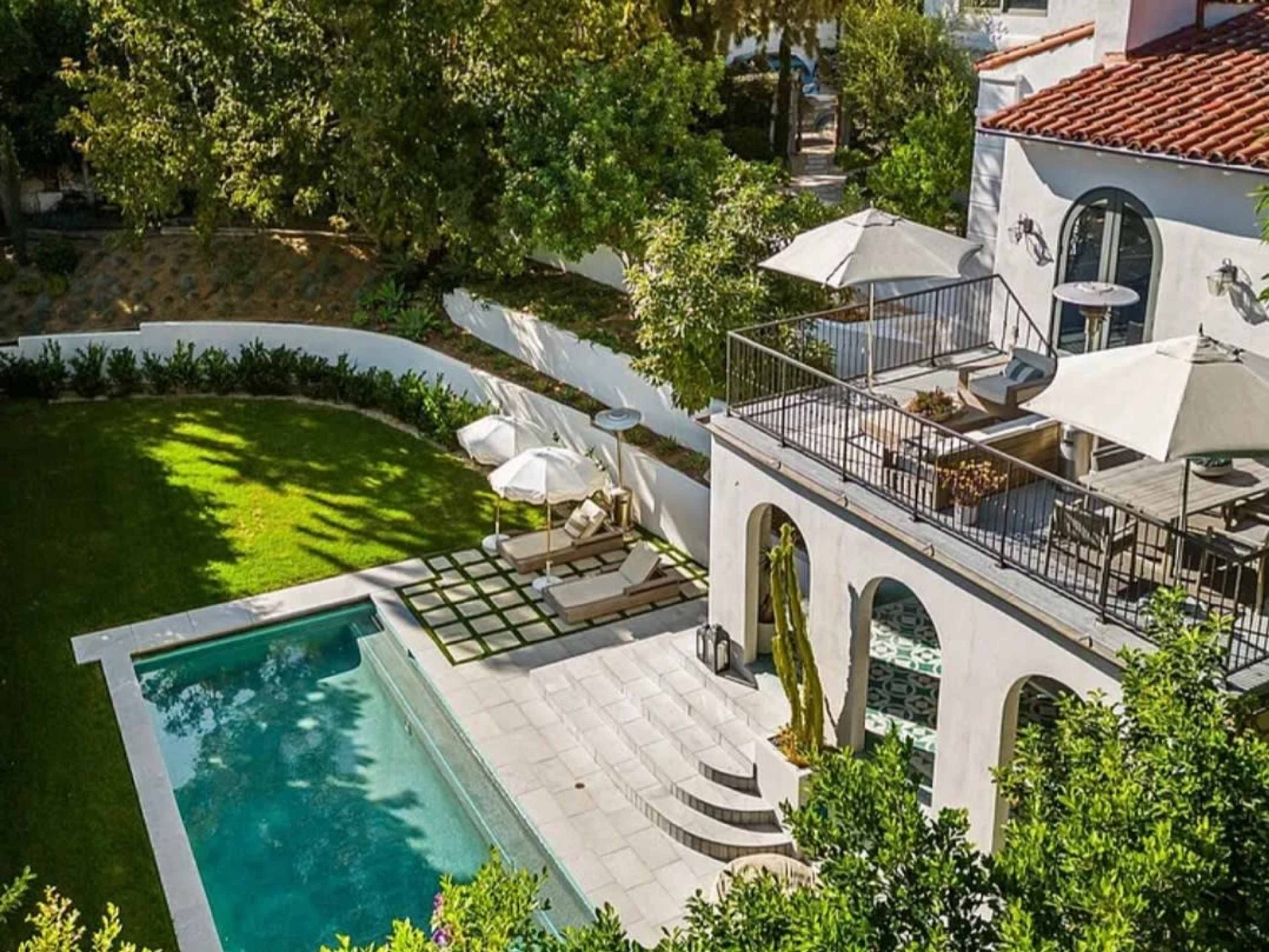 The image shows a spacious backyard featuring a swimming pool, a well-manicured lawn, and a terrace with umbrellas and outdoor furniture.