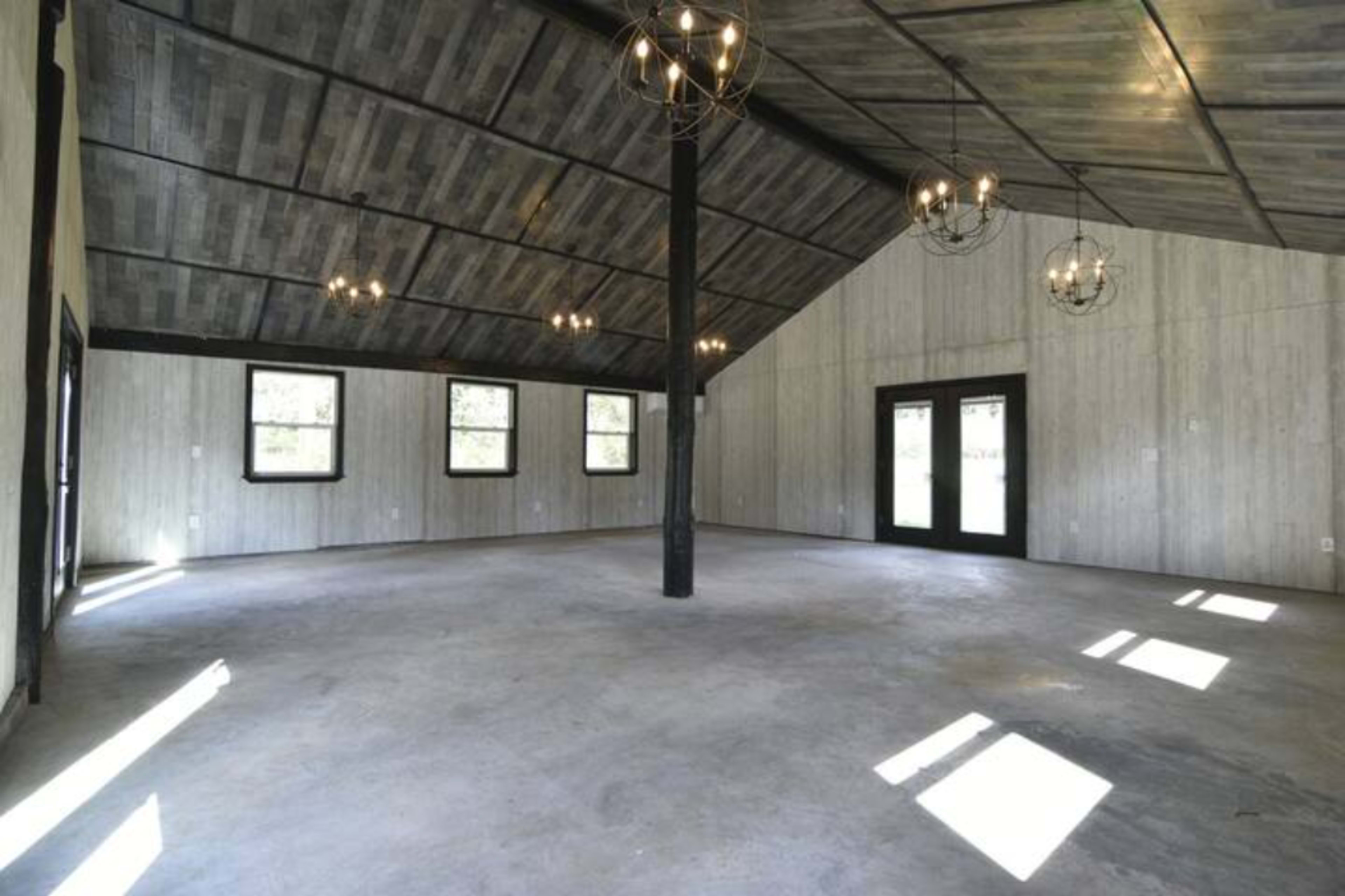 A spacious interior room with a high ceiling, featuring concrete flooring and large windows that let in natural light.