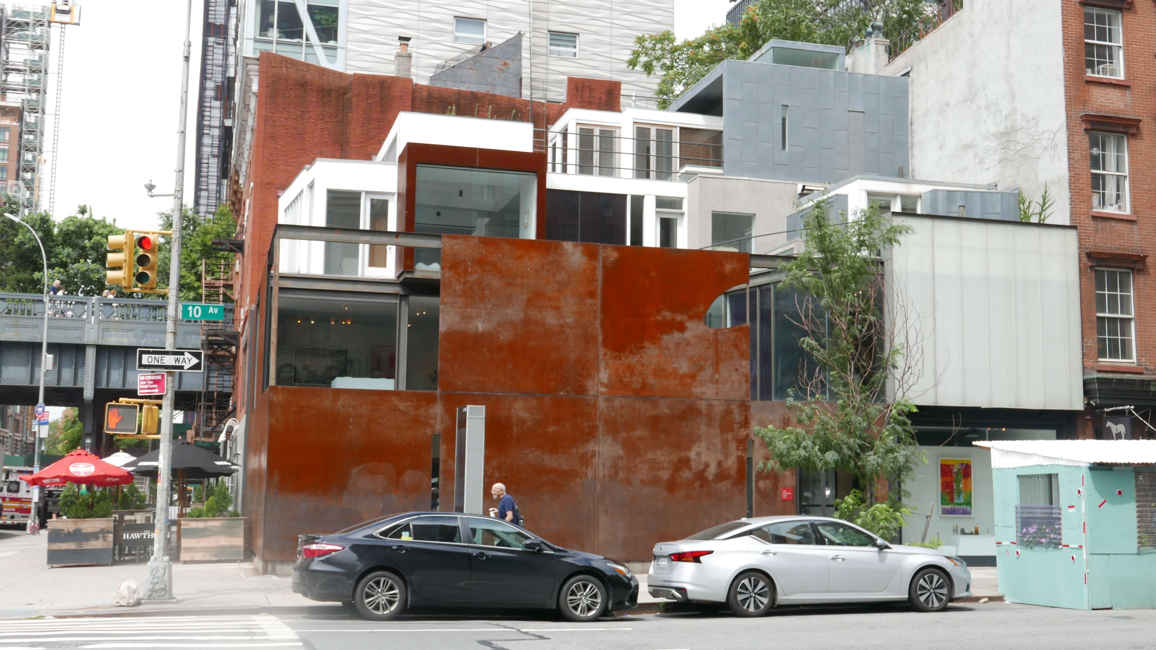 A modern building with a rust-colored metal facade, situated at the corner of a city street with parked cars and a crosswalk.