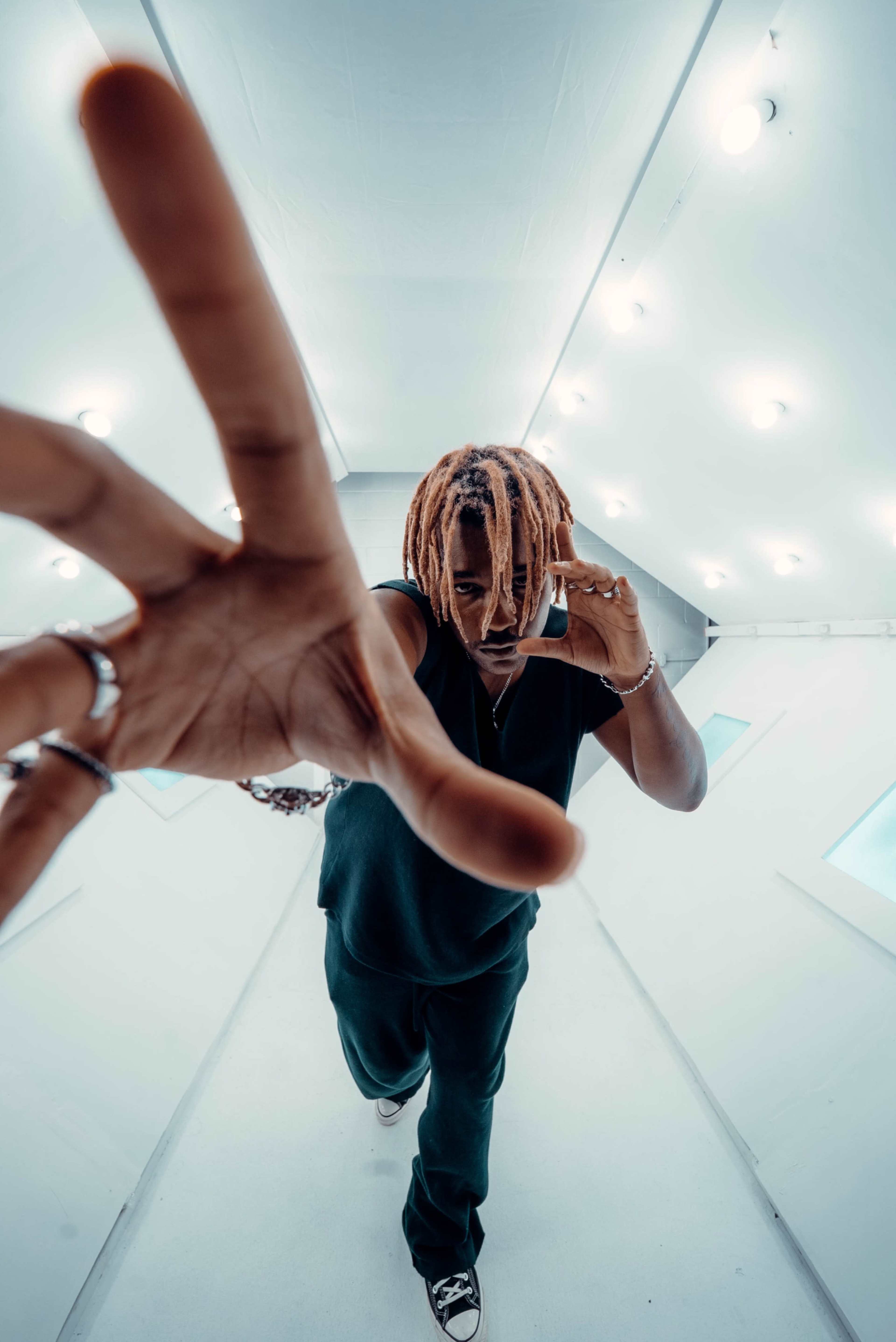 A person with dreadlocks holds a phone to their ear while extending one hand toward the camera in a white, brightly lit space.